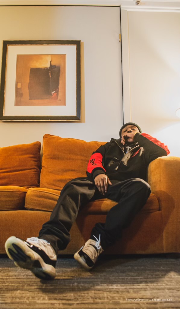 Person sitting on a mustard-colored sofa, resting head on hand, wearing black and red clothing, with artwork on wall behind.
