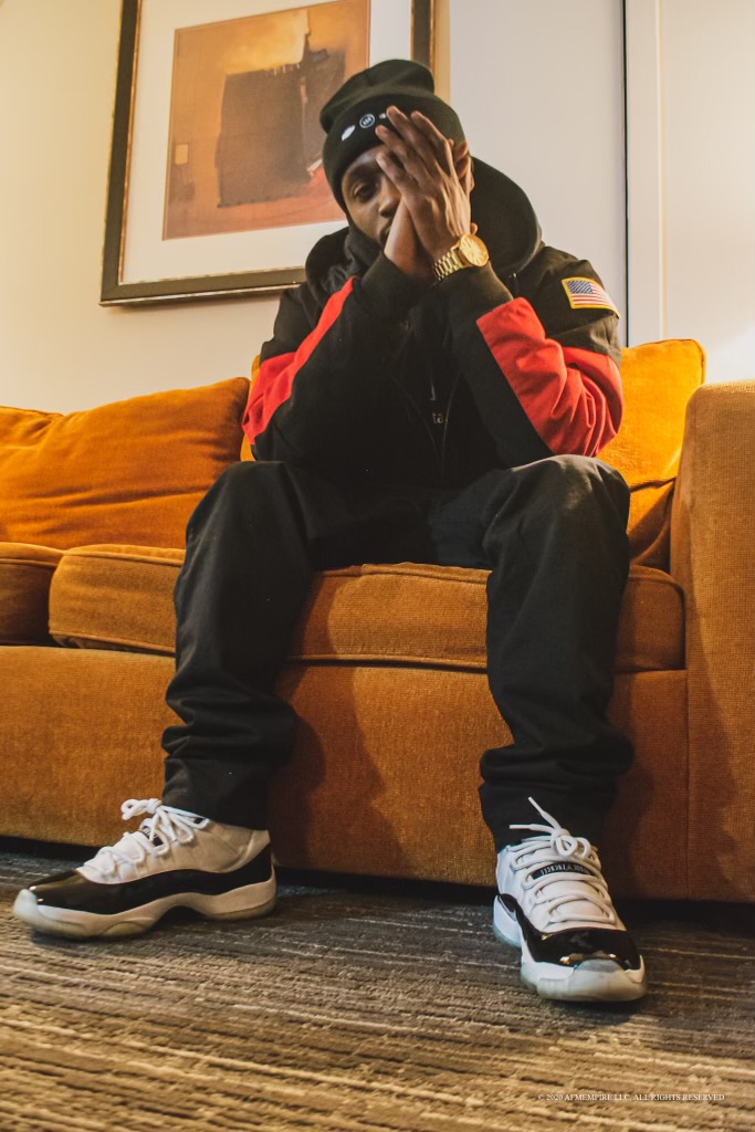 Person sitting on a sofa, wearing a black cap, red and black jacket, and sneakers, taking a photo with a phone.
