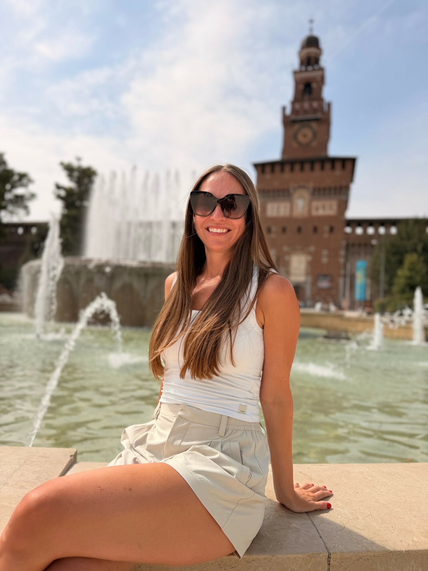 Smiling woman with long hair and sunglasses sitting by a fountain with a historic tower in the background.