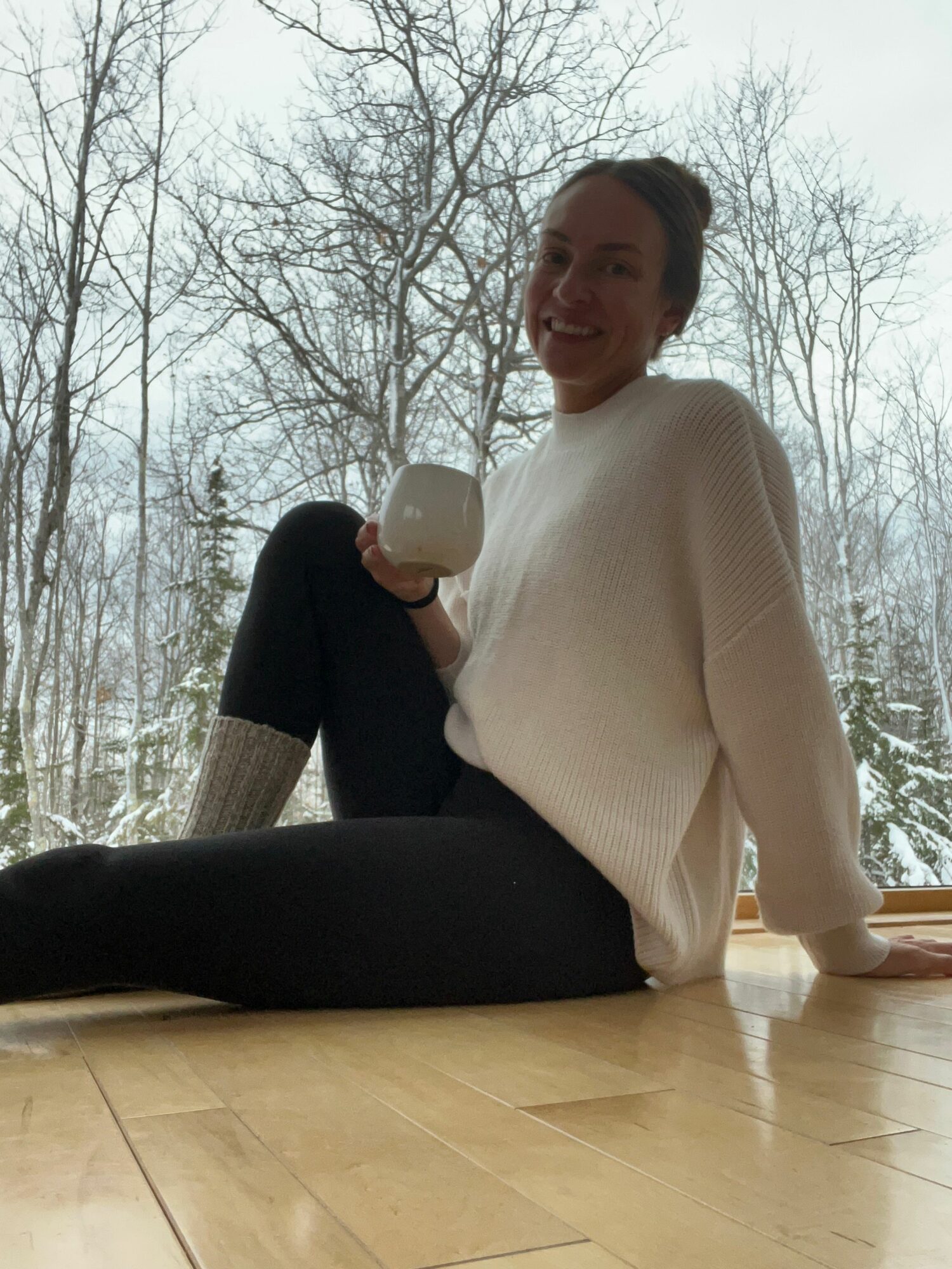 Person kneeling on the floor holding a mug, smiling, with trees visible outside window in background.