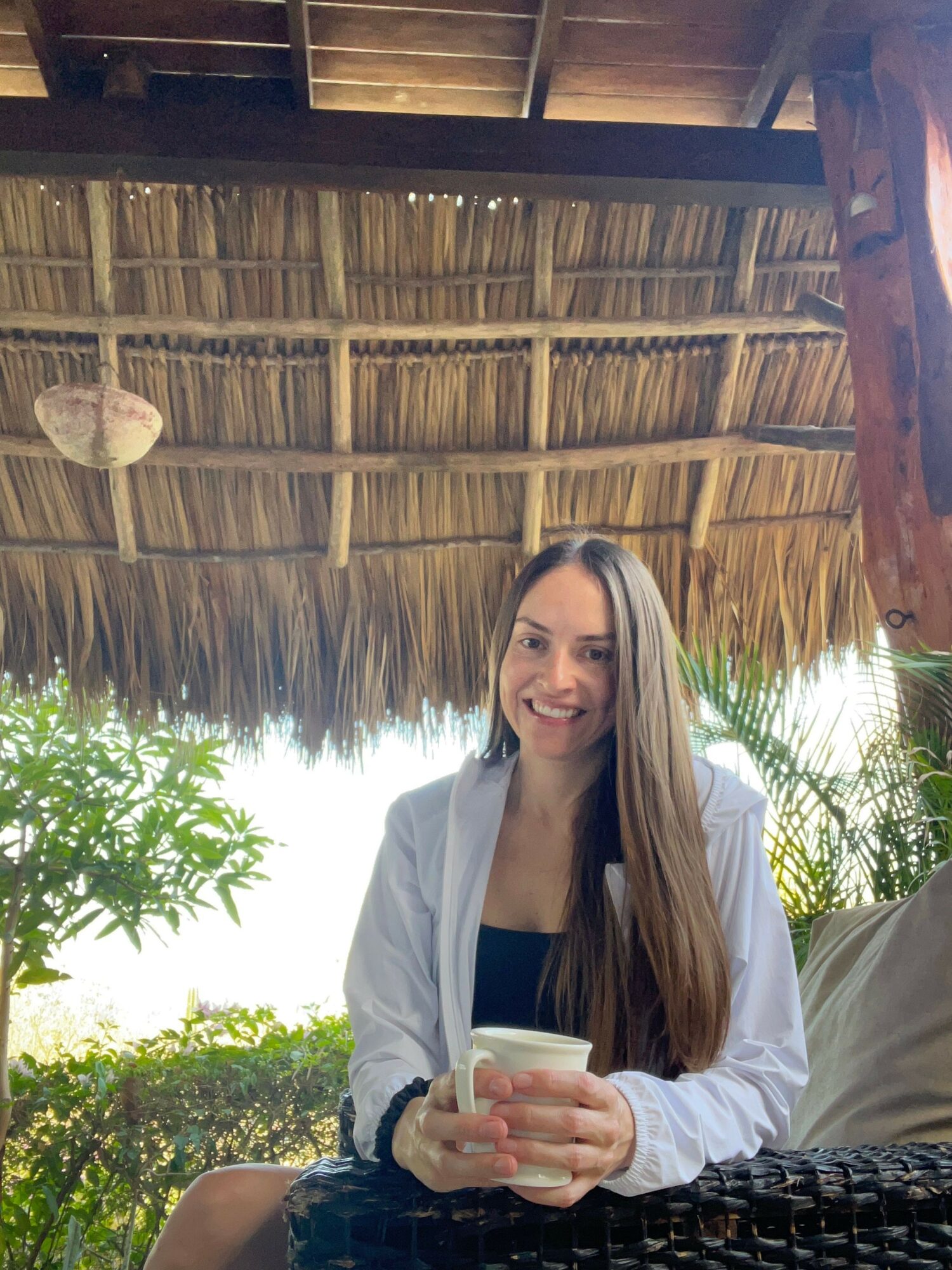 Woman with long hair holding a drink, sitting outdoors with a bamboo fence and greenery in background.
