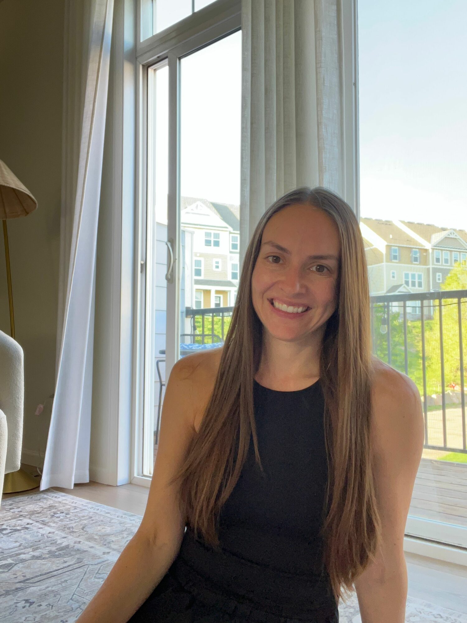 Young woman with long hair smiling, sitting indoors near large windows with a view of buildings and a balcony, sunlight streaming in.