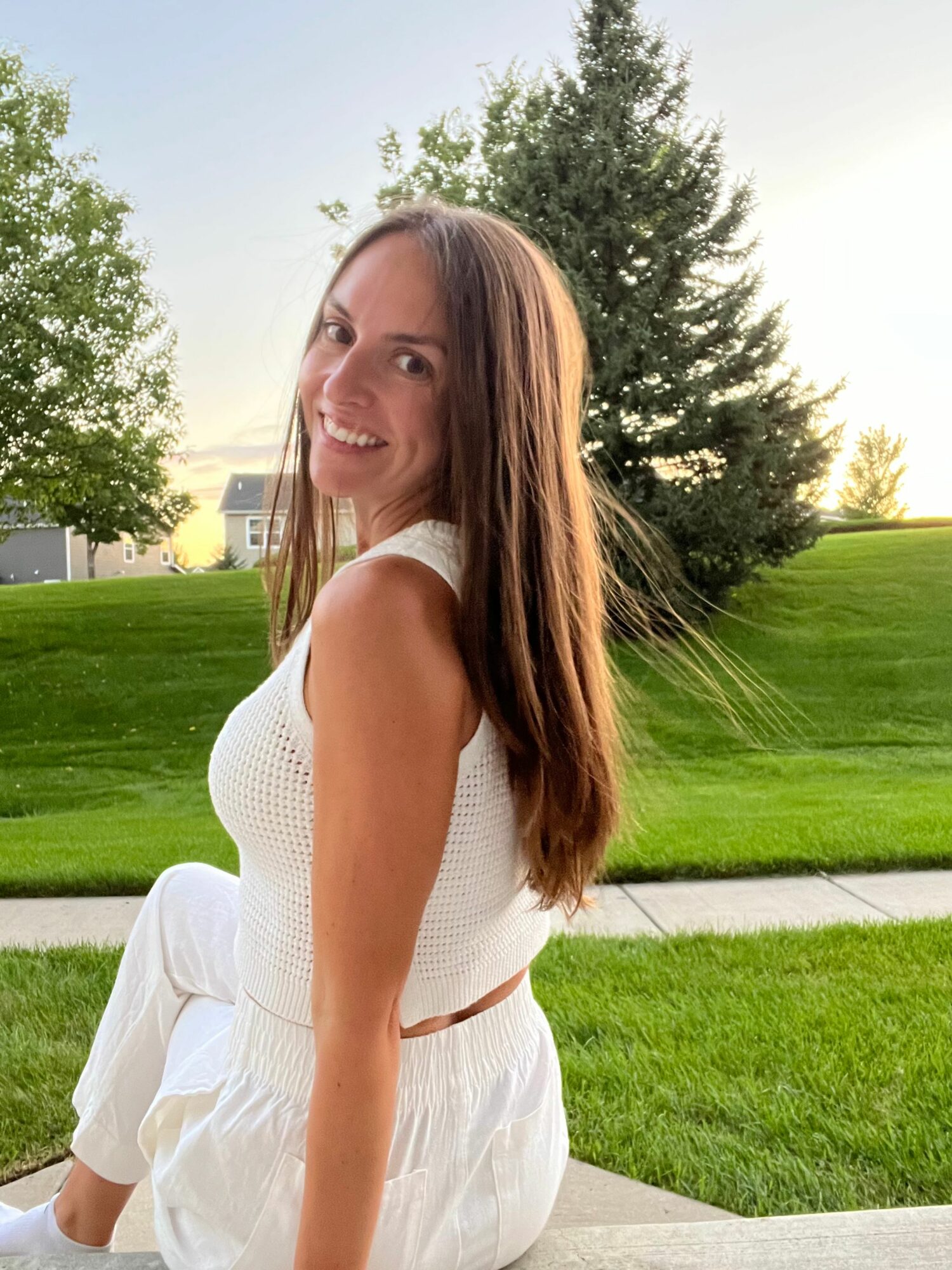 Young woman with long hair smiling outdoors in a park, wearing a white dress, with trees and grass in the background.