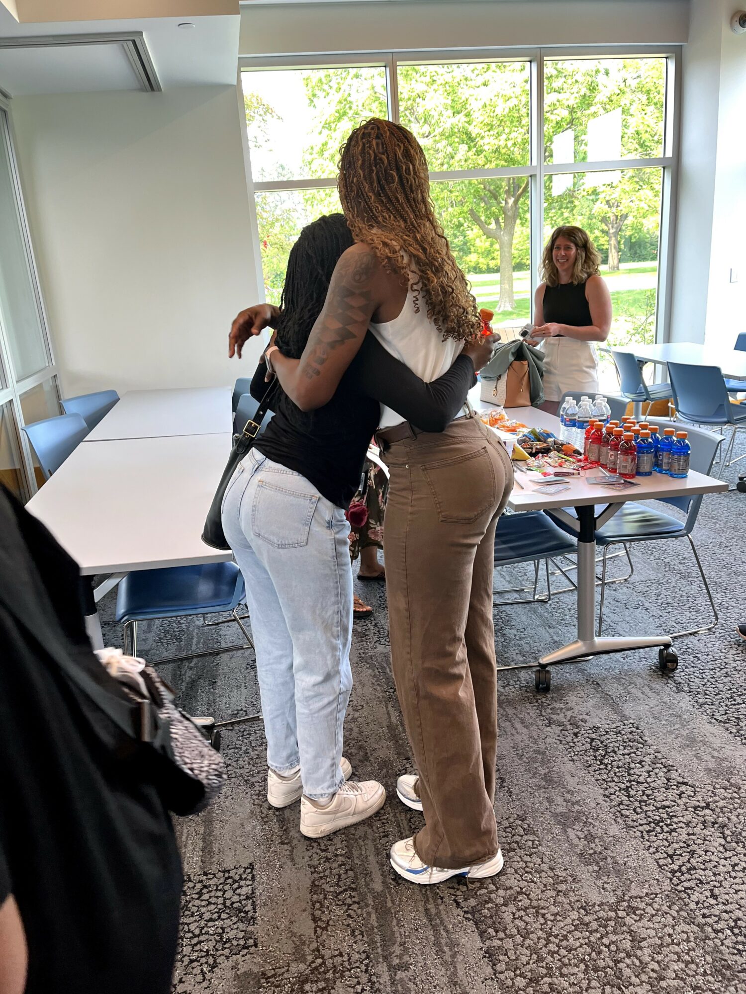 Two women hugging in a room with large windows, tables, and chairs, with a woman sitting at a table in the background.