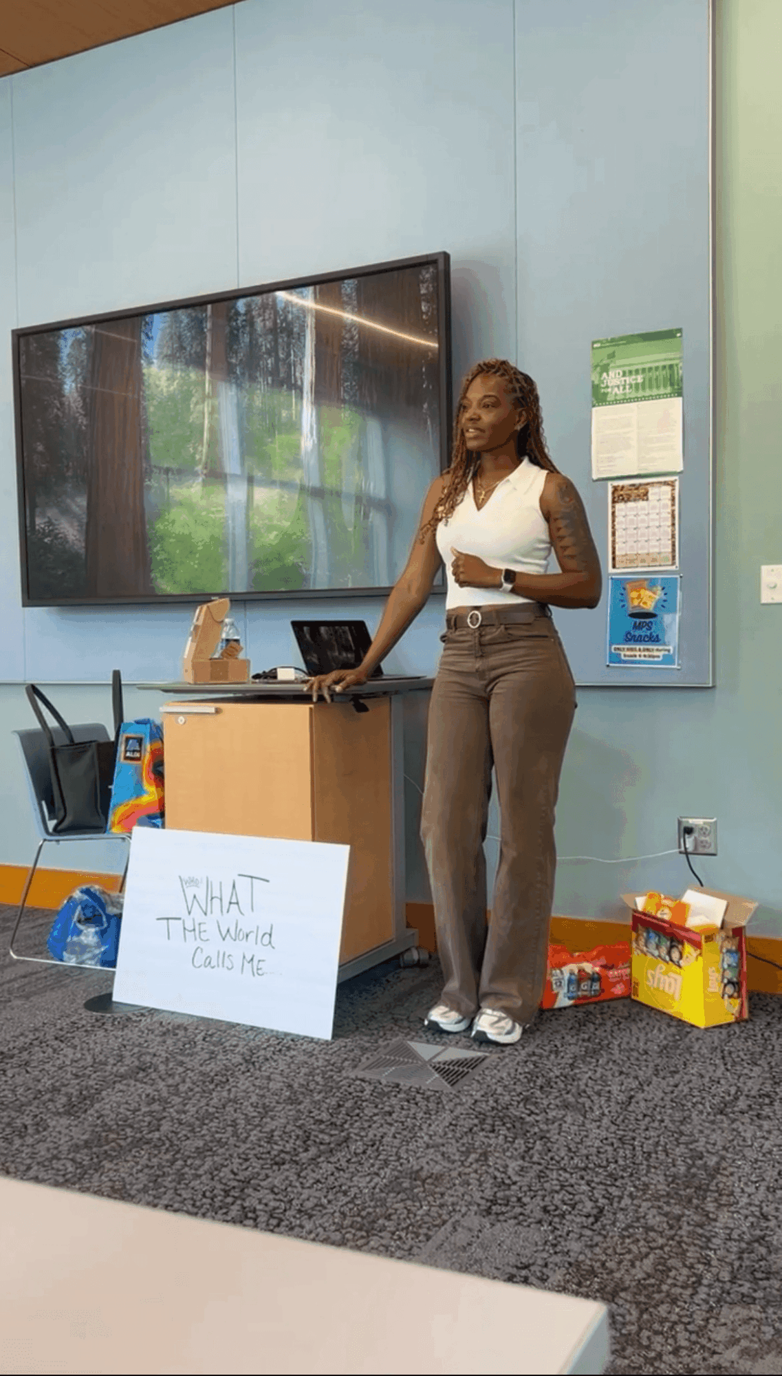 Woman standing next to a table with a sign, in a room with a large screen and colorful boxes on the floor.