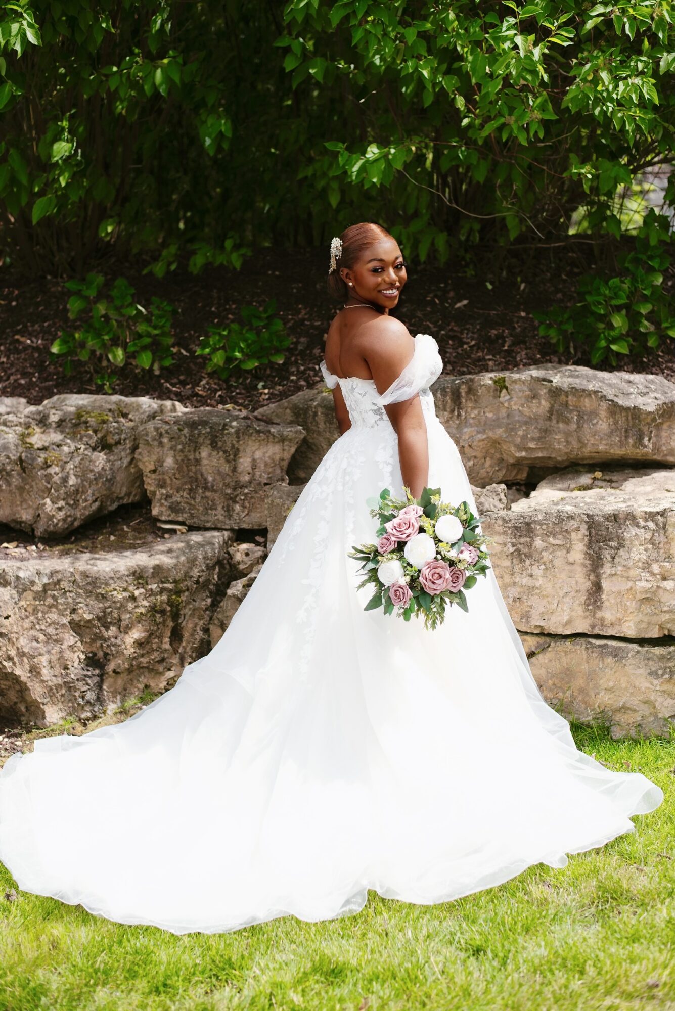 Bride in a white wedding dress holding a bouquet, standing outdoors in front of rocks and greenery.