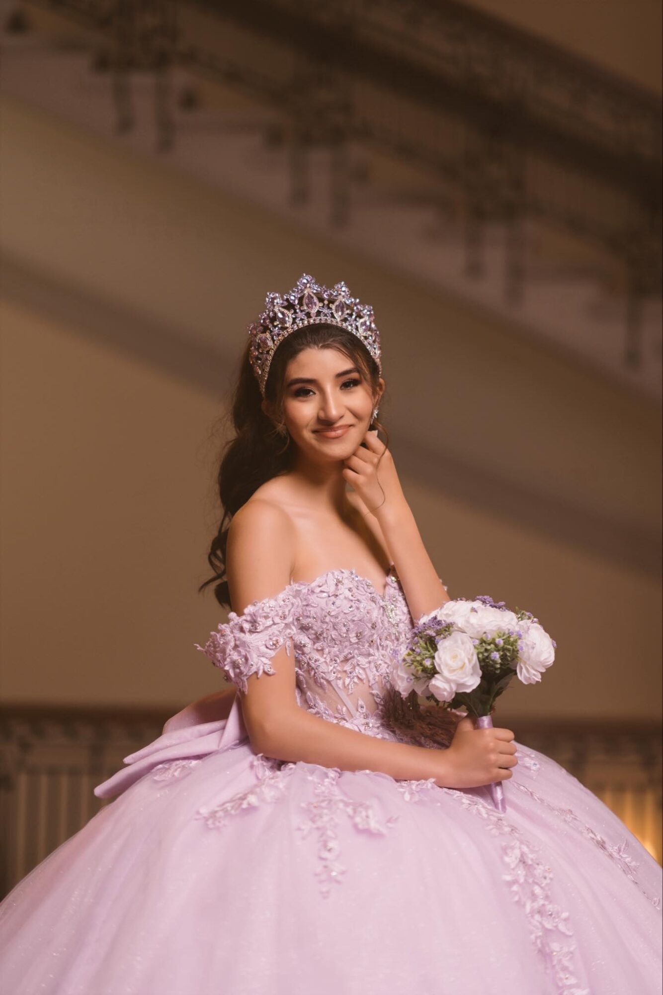 Young woman in a pink ball gown with floral details, wearing a tiara, holding a bouquet of white flowers, smiling.