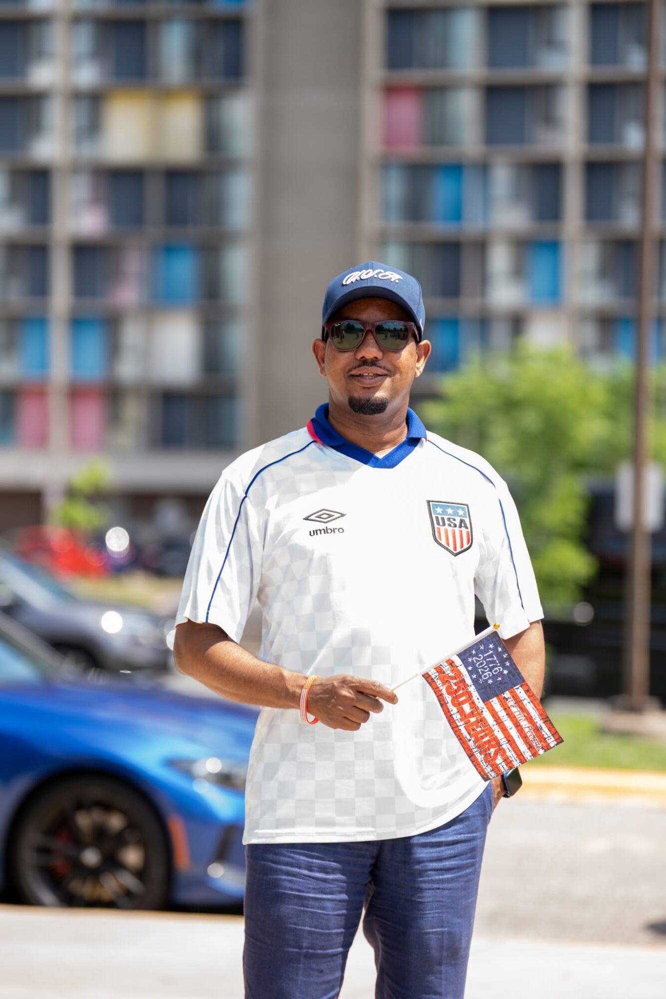 Man wearing sunglasses, a cap, and a USA soccer jersey holding a small American flag outdoors.