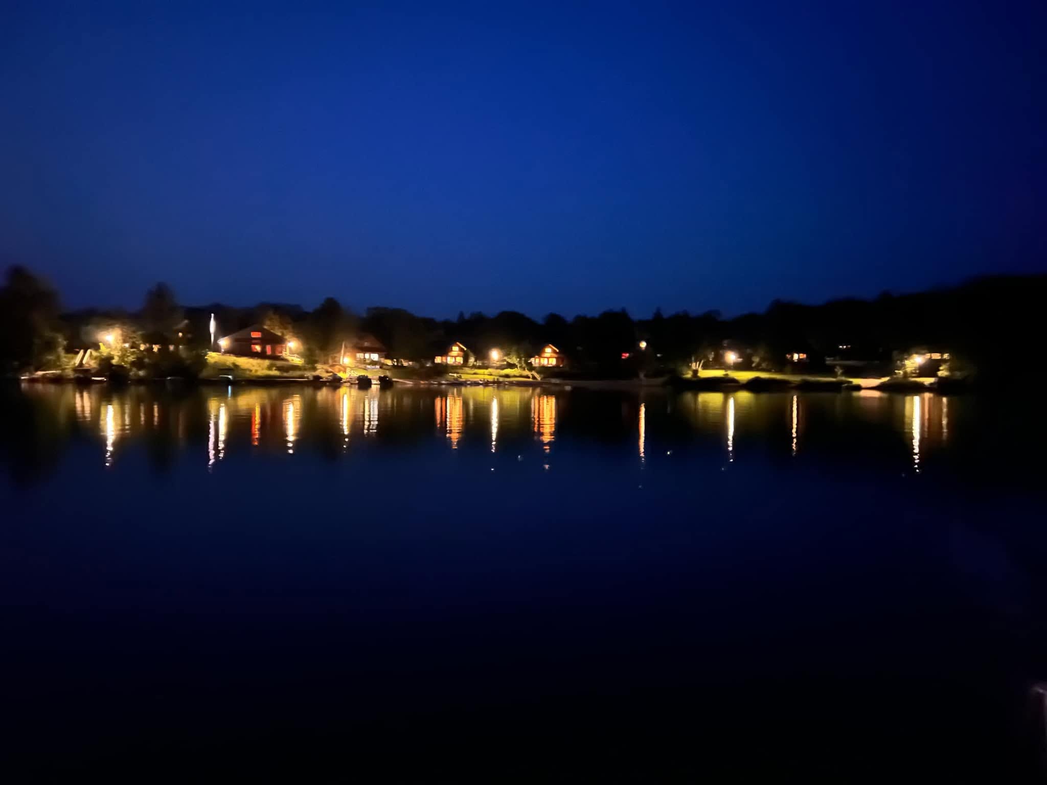 Nighttime scene of a calm body of water reflecting lights from houses along the shoreline.
