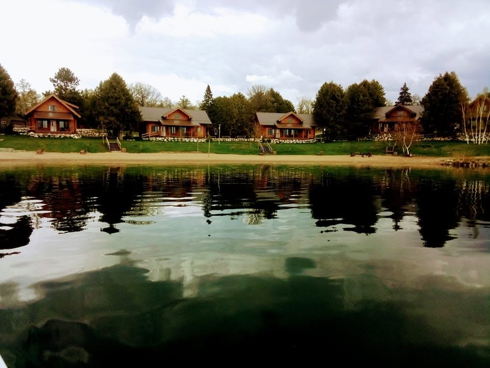 Houses along a lakeshore with trees and cloudy sky, reflected in the water, landscape scene.