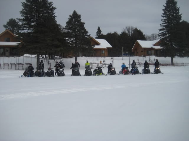 Group of people on snowmobiles in snowy landscape with trees and houses in background.