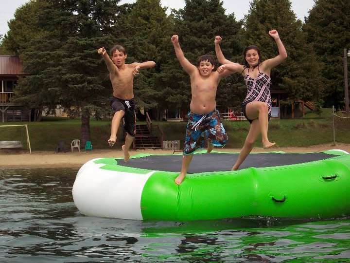 Three children jumping on an inflatable raft near water, with trees and houses in the background.