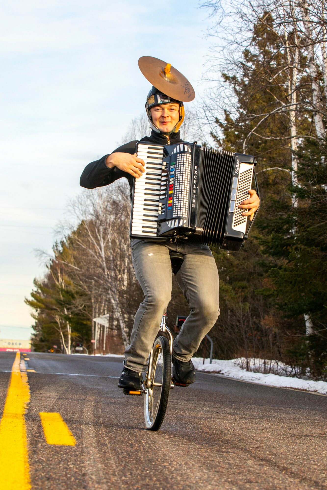 Person riding a unicycle on a road, playing an accordion, wearing a hat with a large object on top, trees in background.