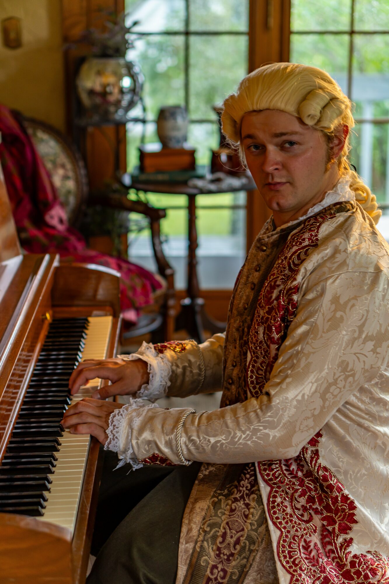 Person dressed in historical costume playing piano in a room with large window and decorative furniture
