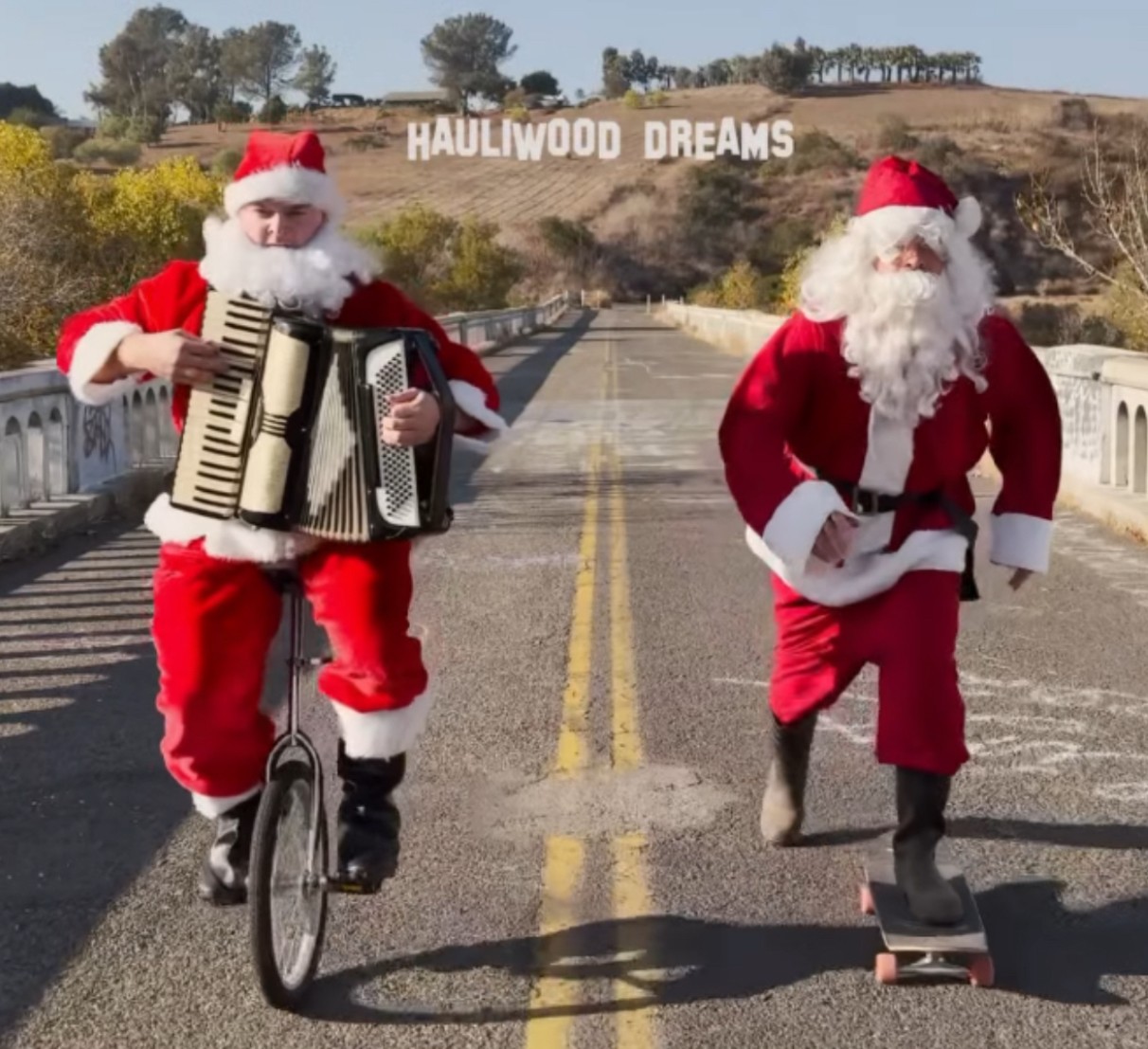 Two people dressed as Santa Claus, one playing an accordion and riding a bicycle, the other skateboarding, on a road with a sign reading 'Haulwood Dreams' in the background.