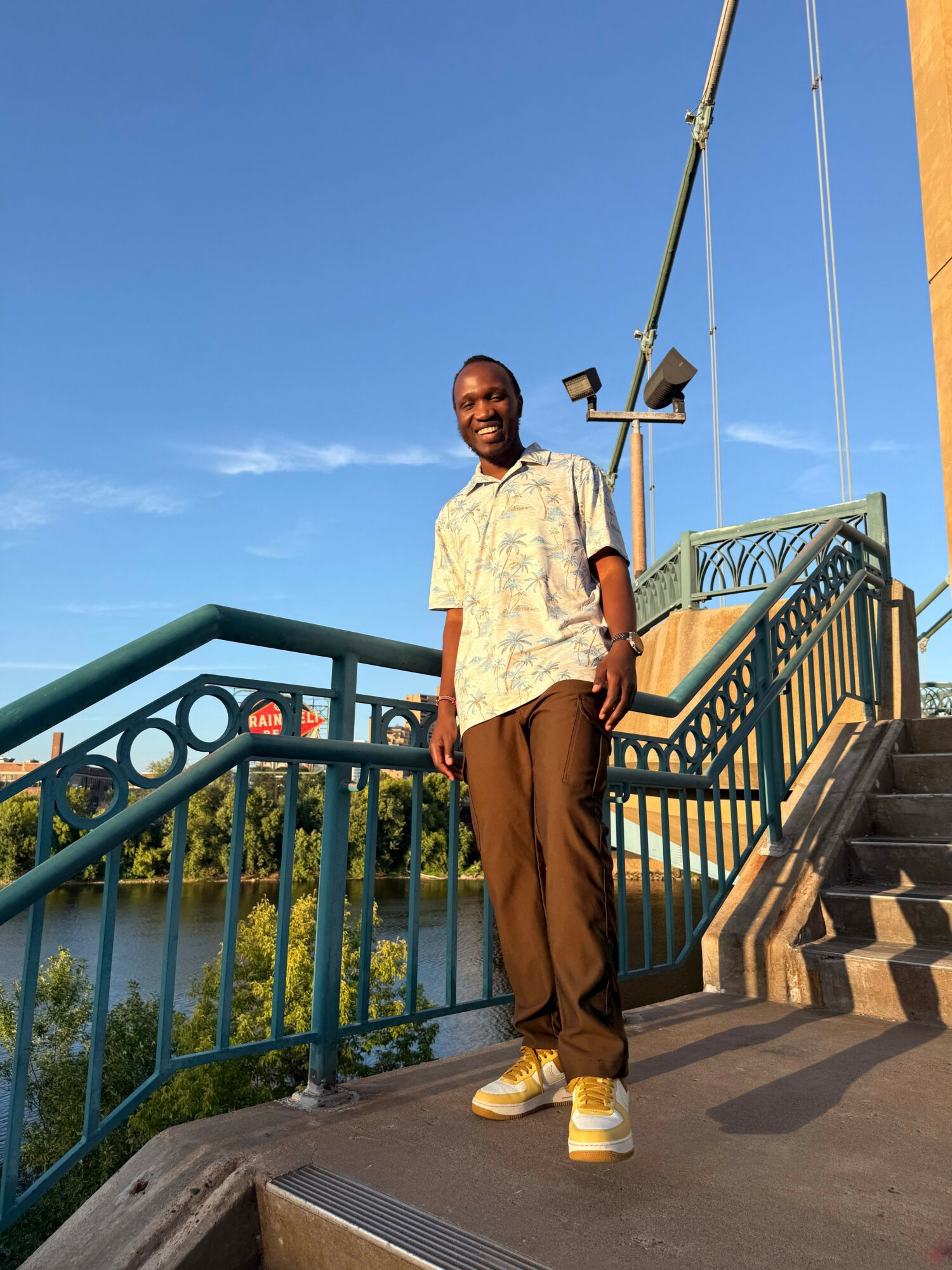 Man standing on sidewalk near green railing and stairs, with blue sky and clouds in background.