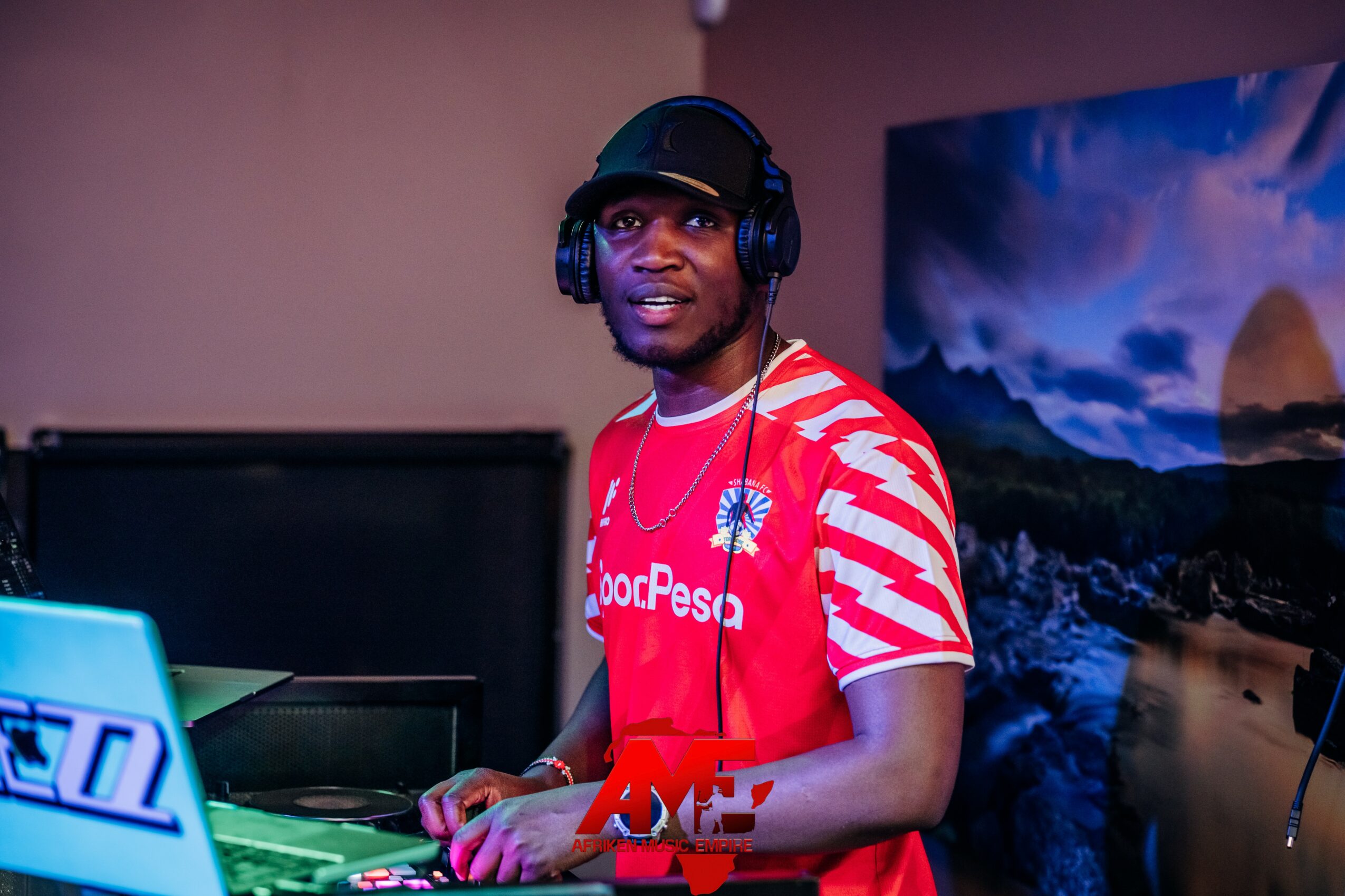 Young man wearing headphones and a red and white striped shirt, standing in front of a computer and a scenic backdrop.