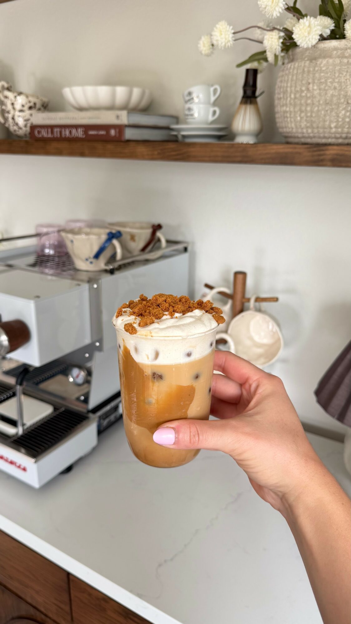 Hand holding a glass of iced coffee with whipped cream and caramel drizzle, background includes a shelf with flowers and kitchen items.