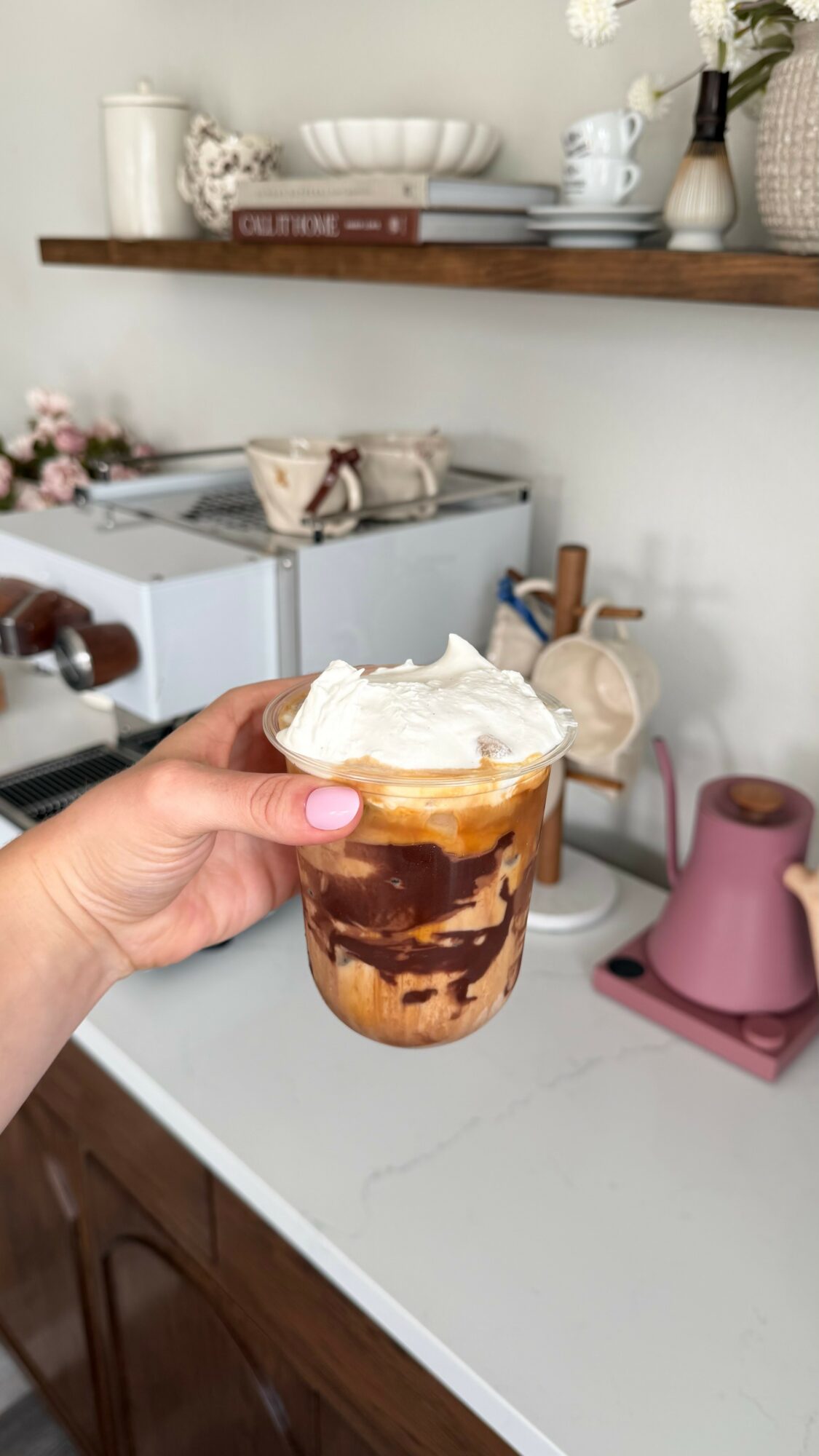 Hand holding a glass of iced coffee with whipped cream, chocolate syrup, and ice cubes, in a kitchen setting.