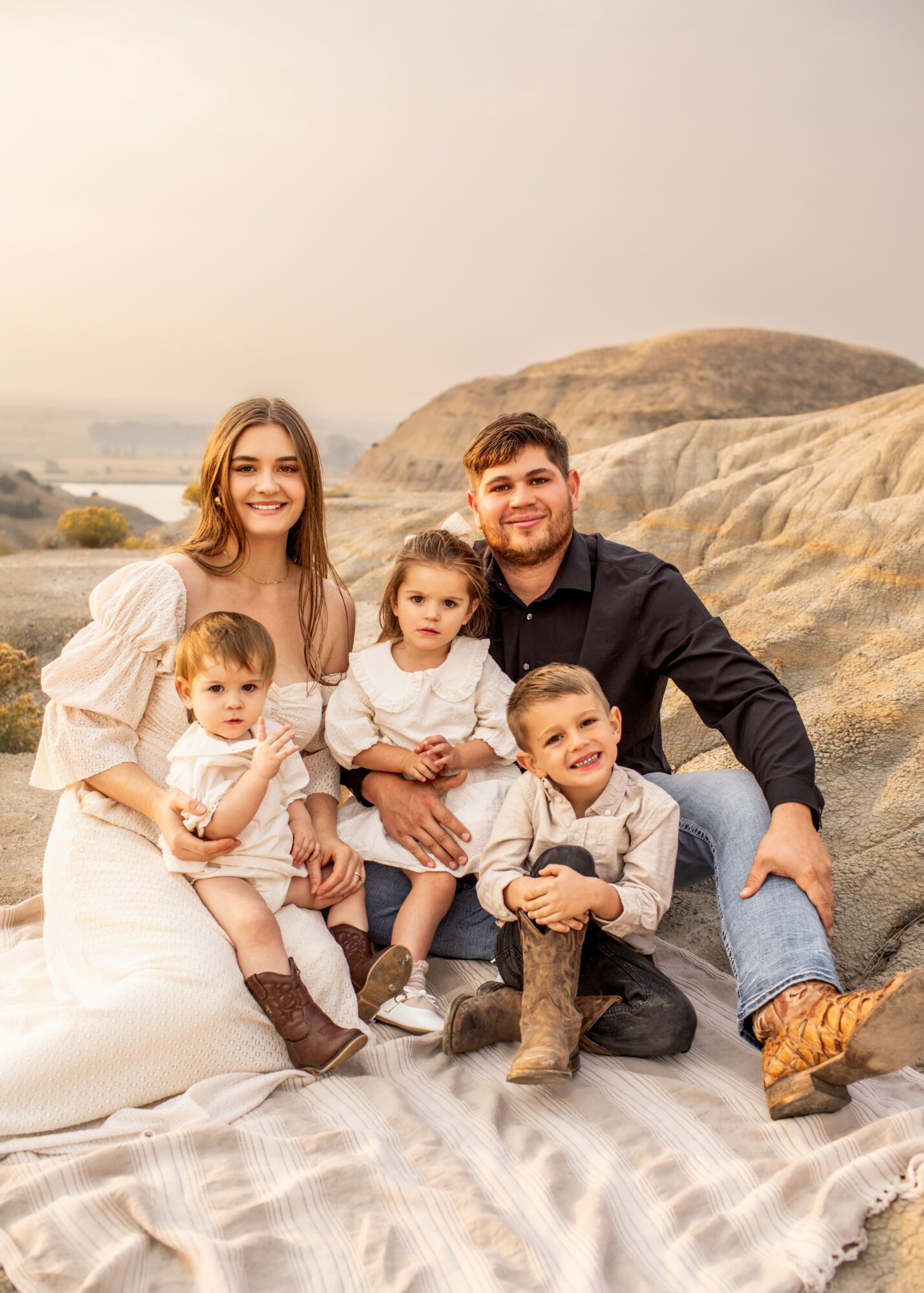 Family of five sitting outdoors on a blanket with rocks and hills in the background, smiling at the camera.