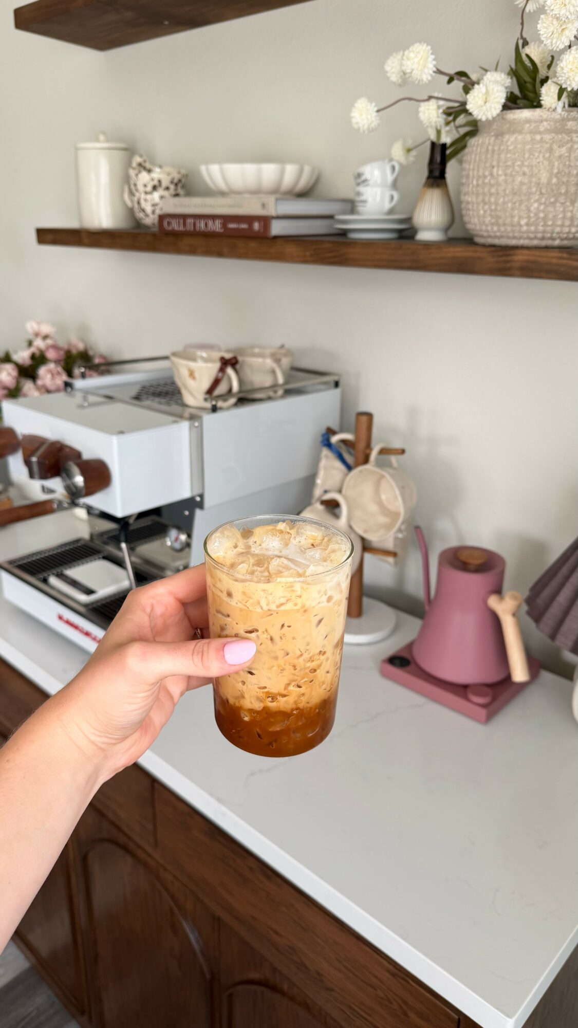 Hand holding a glass with a layered beverage on a kitchen counter, with shelves and decorative items in the background.