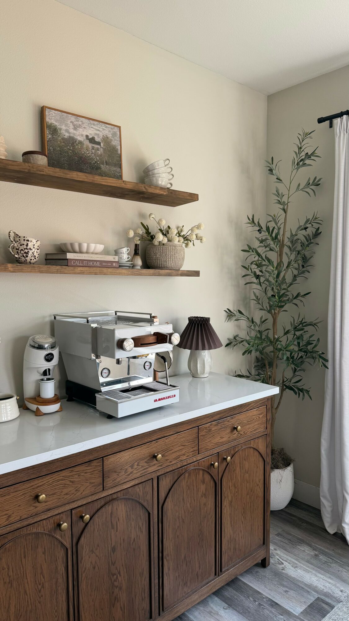 Wooden cabinet with white countertop, coffee machine, lamp, and decorative items in a room with shelves and a plant.