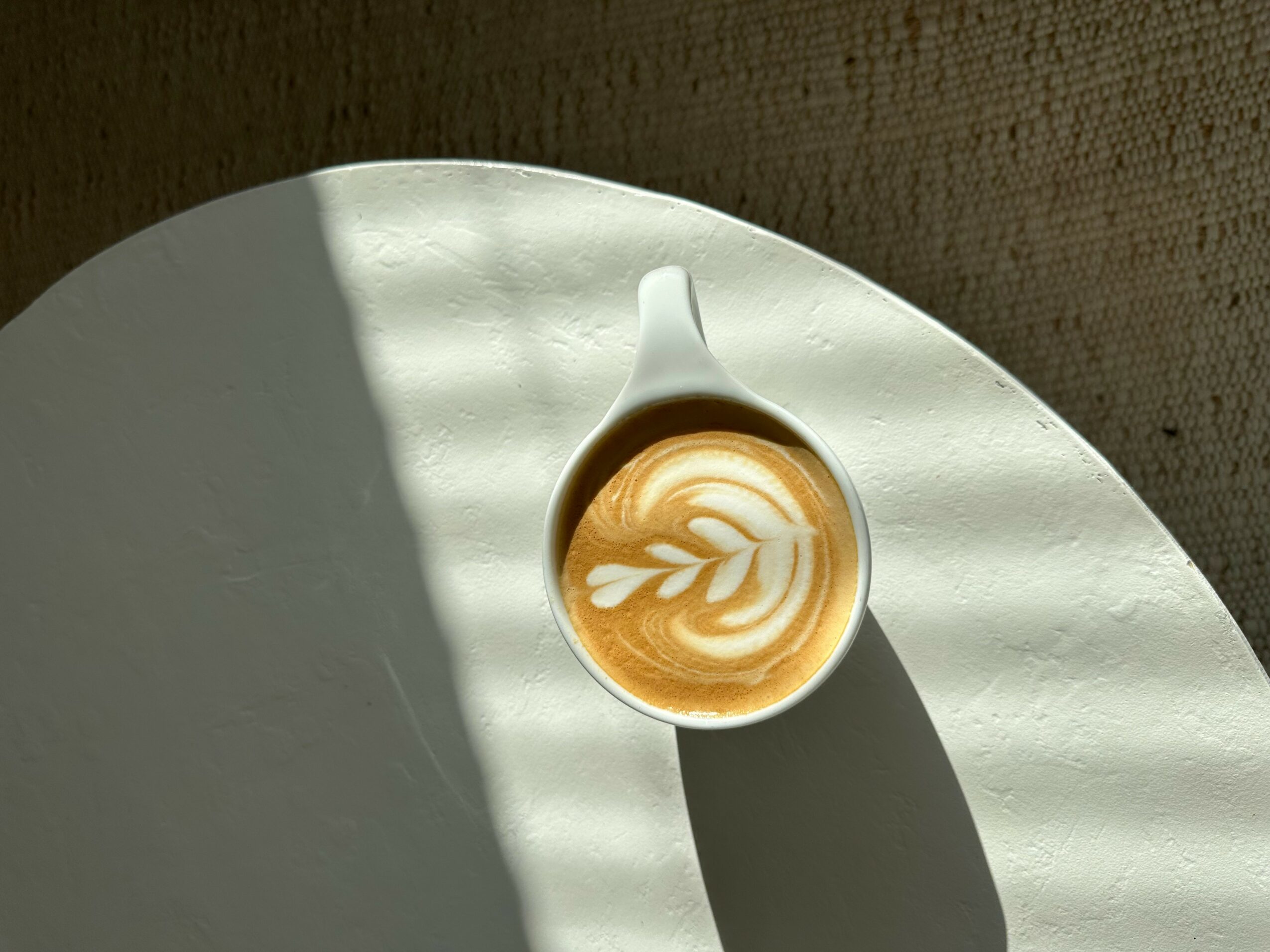 A cup of coffee with latte art on a white table, shadow cast across the surface.