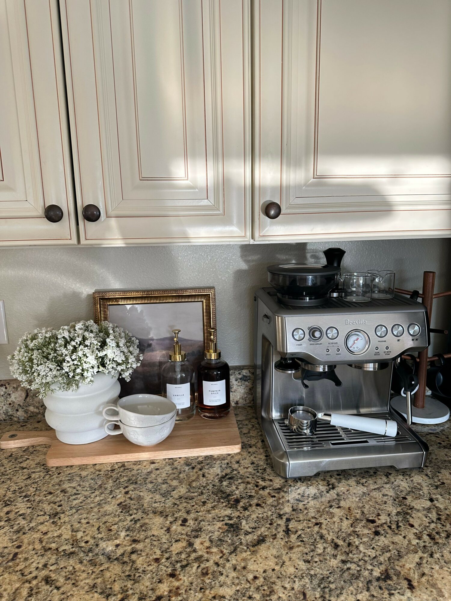Coffee machine, bottles, cups, and flowers on a kitchen countertop with cream-colored cabinets in the background.