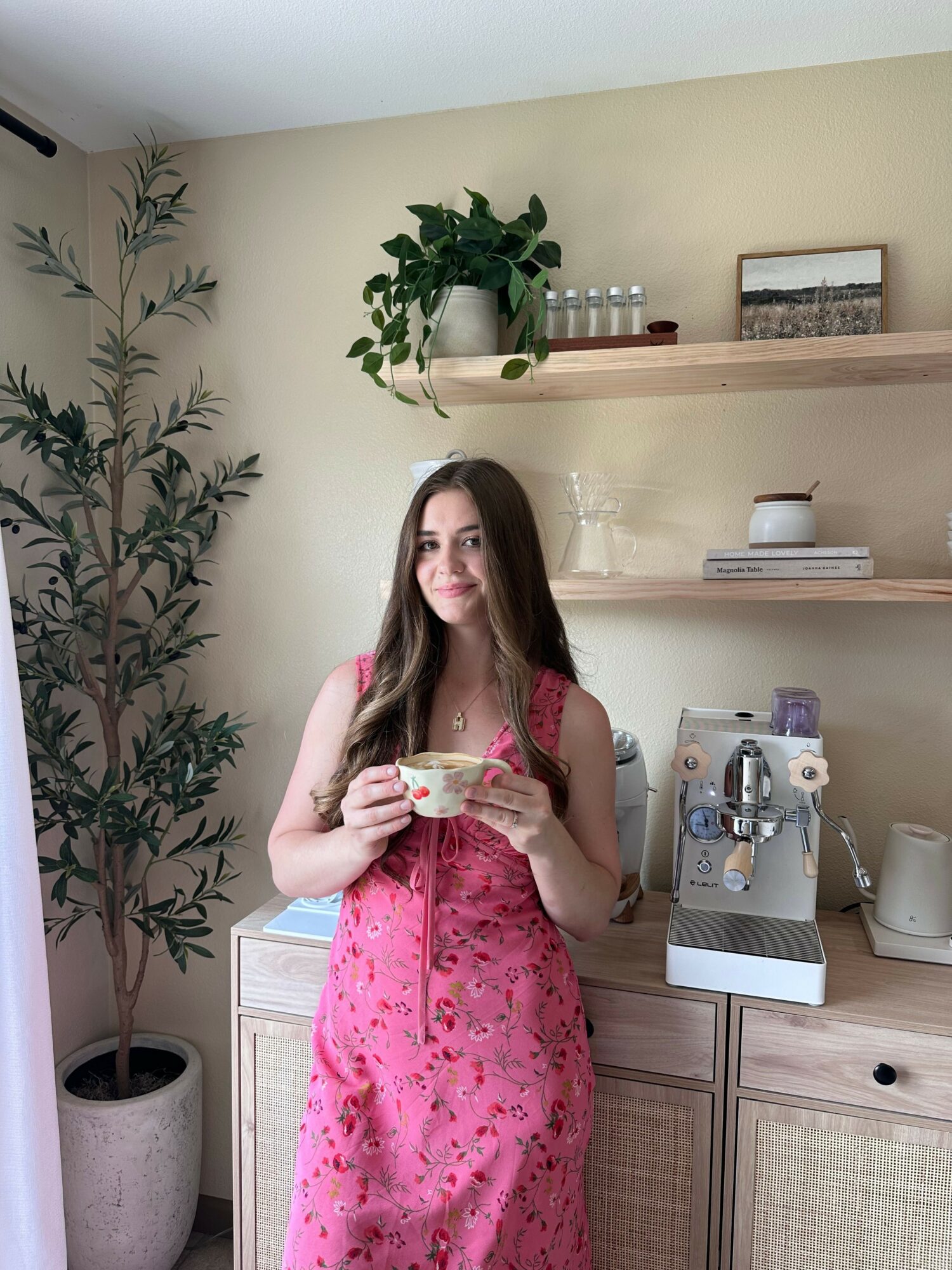 Woman in pink floral dress taking mirror selfie in a room with plants and furniture.
