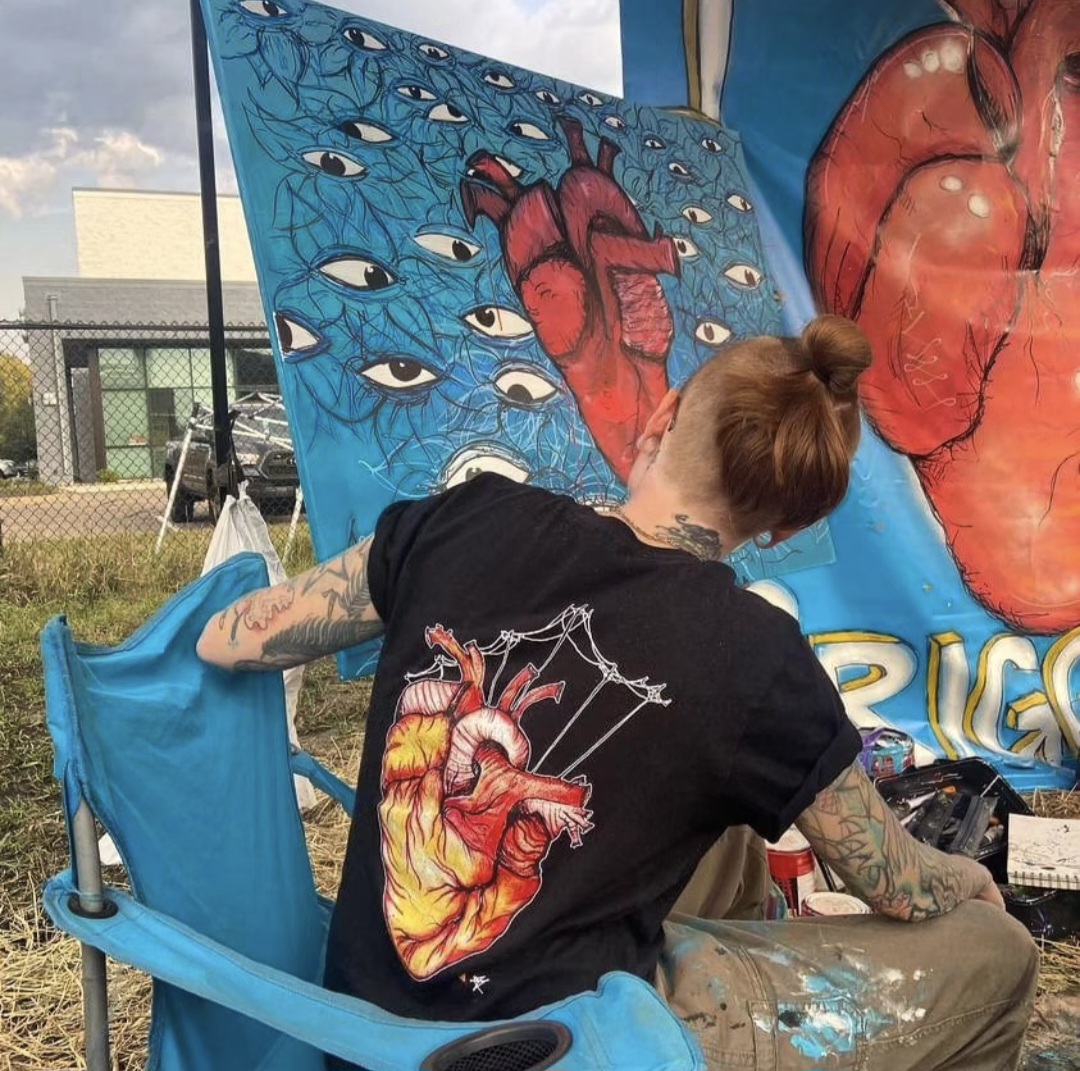Person painting colorful heart and eye artwork on large canvas outdoors, with art supplies nearby, wearing a black T-shirt with a heart design.