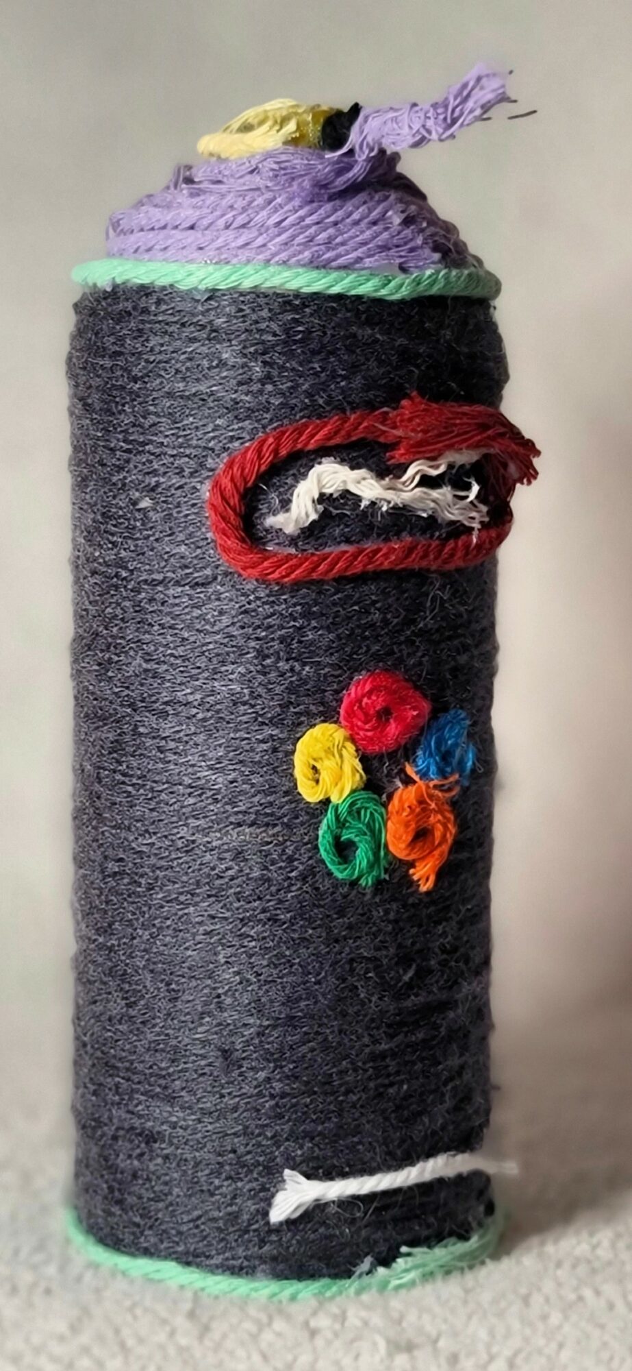 A cylindrical object wrapped in dark gray fabric with colorful yarn decorations, including a red mouth and multicolored flowers, against a plain background.