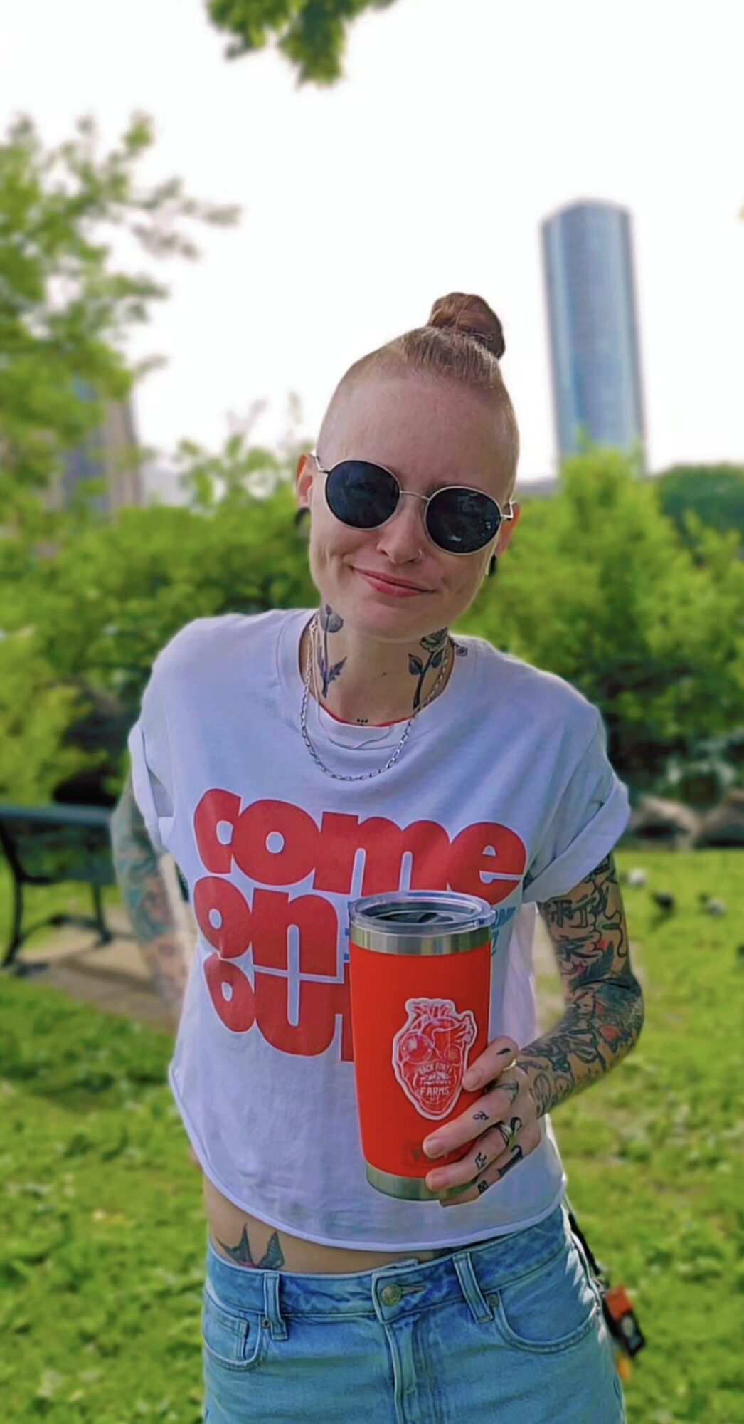 Person with sunglasses and tattoos holding a red cup outdoors, smiling, with trees and a tall building in the background.