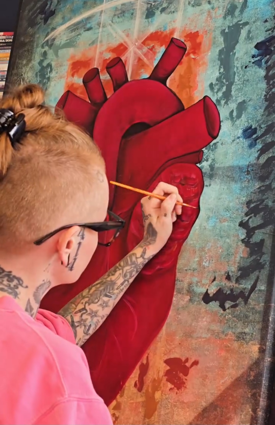 Person painting a large red anatomical heart on a colorful wall, with tattoos and glasses, using a small brush.