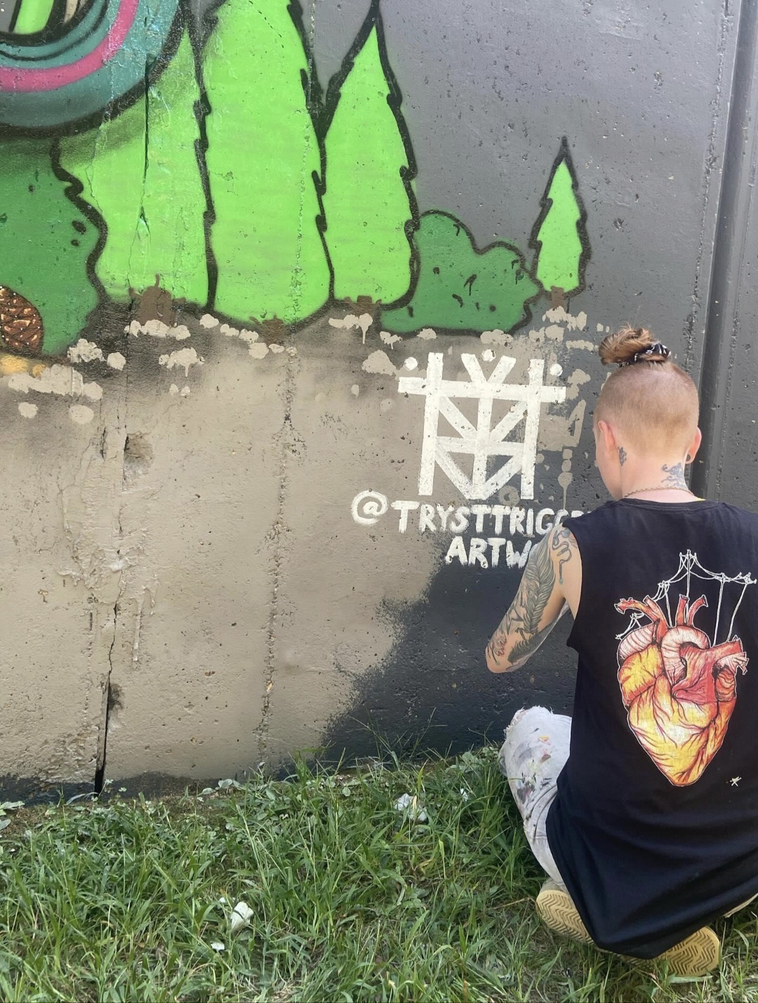 Person with a tattooed arm and a bun hairstyle crouches in front of a graffiti mural on a concrete wall, with green trees painted above.