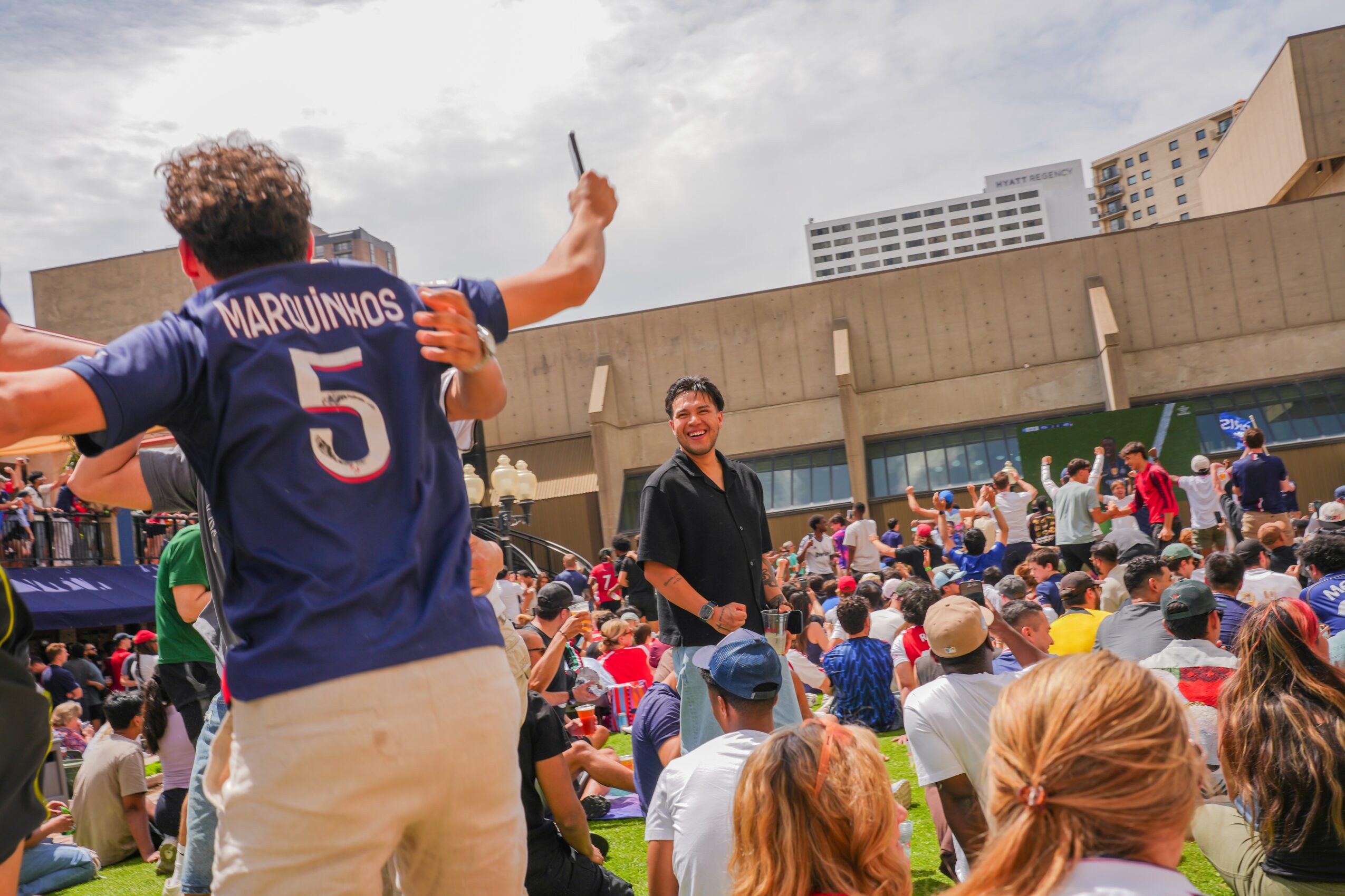 Crowd gathered outdoors with a person standing on a platform, holding a microphone, addressing the audience, with buildings in the background.