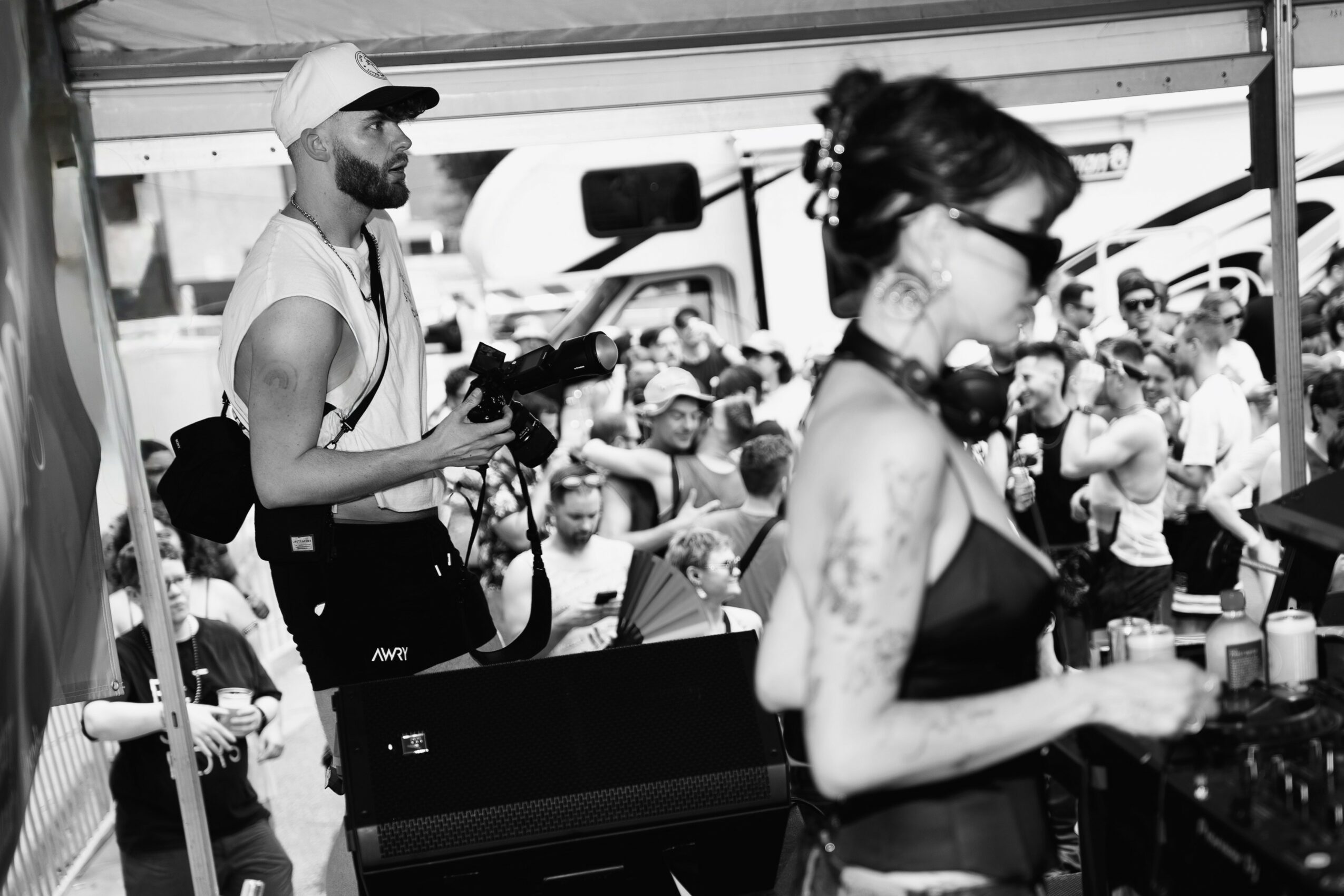 A woman with tattoos and sunglasses stands at a DJ booth, with a crowd of people in the background.