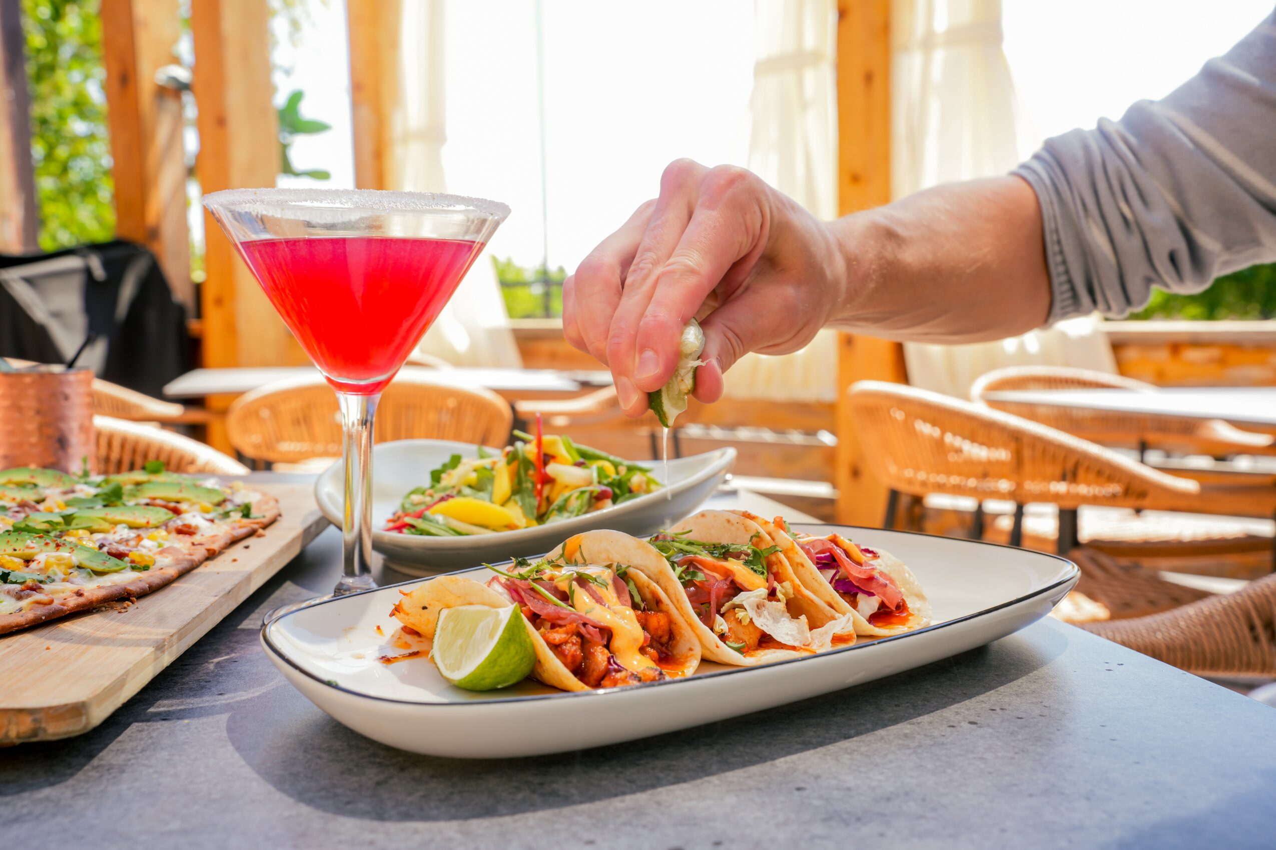 Person's hand adding toppings to tacos on a white plate, with a cocktail and pizza in the background, in a bright room.