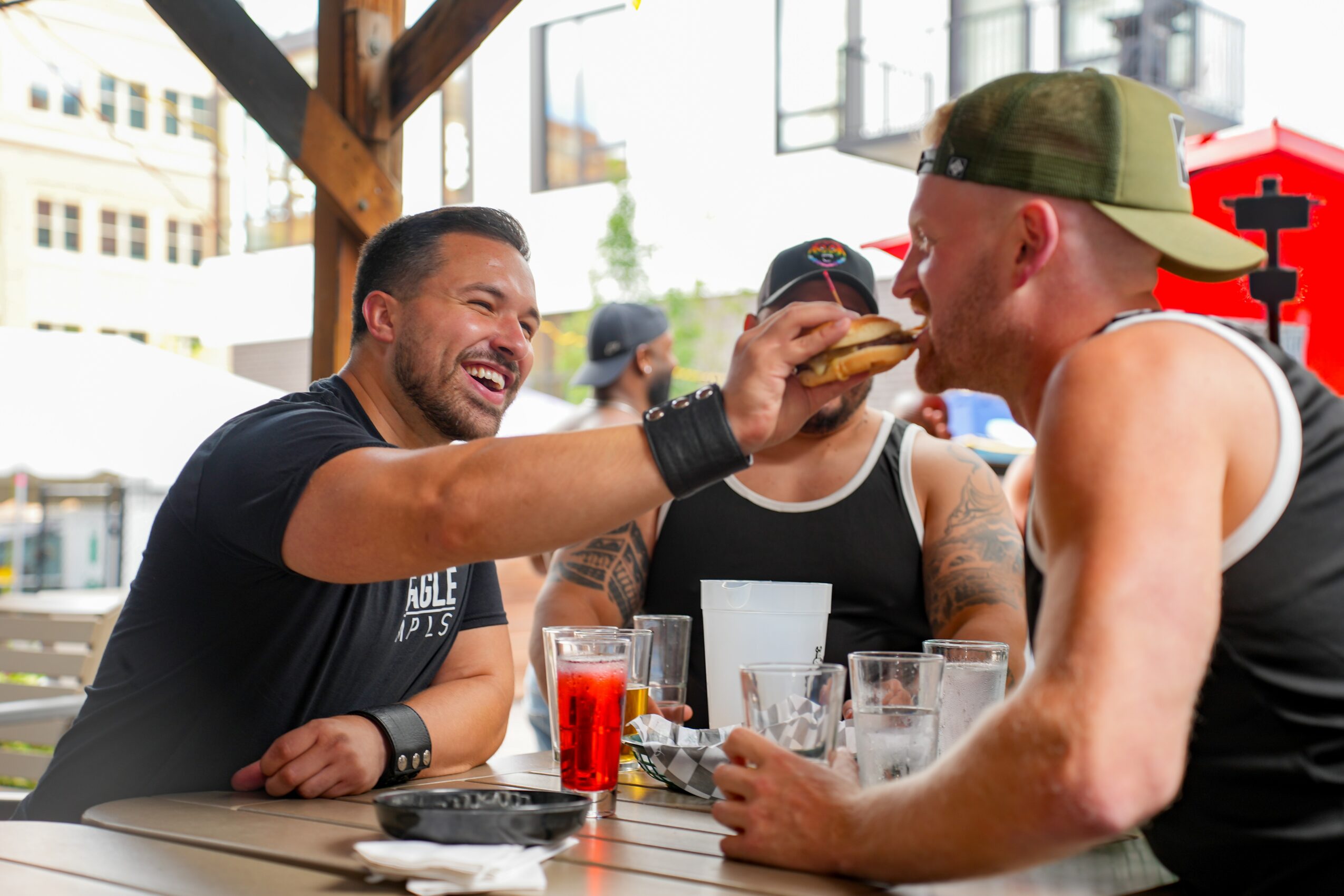 Three men sitting at an outdoor table, one holding a burger near another's mouth, smiling and enjoying drinks.