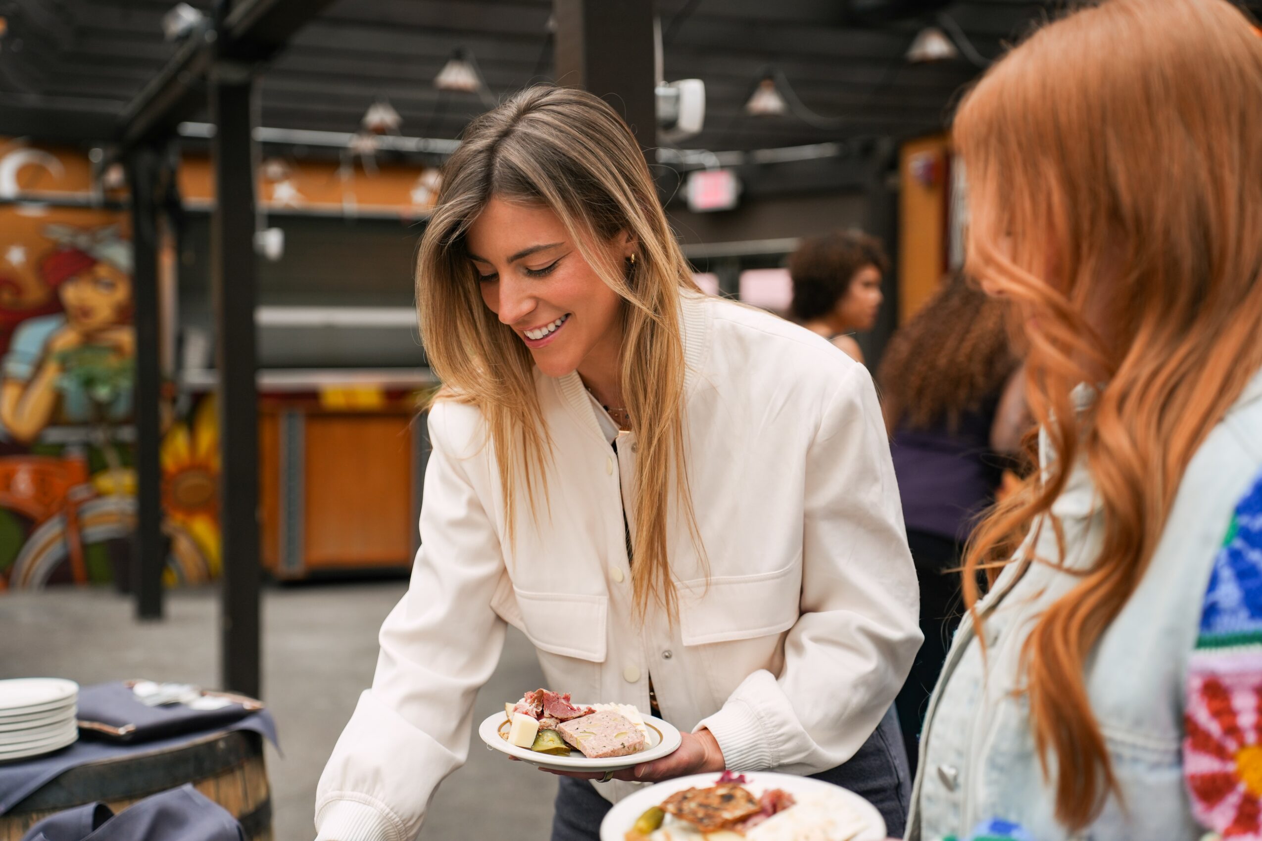 Two women smiling and serving food at a social gathering indoors, with plates and a table in the foreground.
