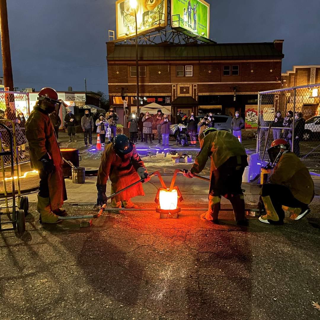 People gather around a glowing object on the ground at night, with a fence and building in the background.