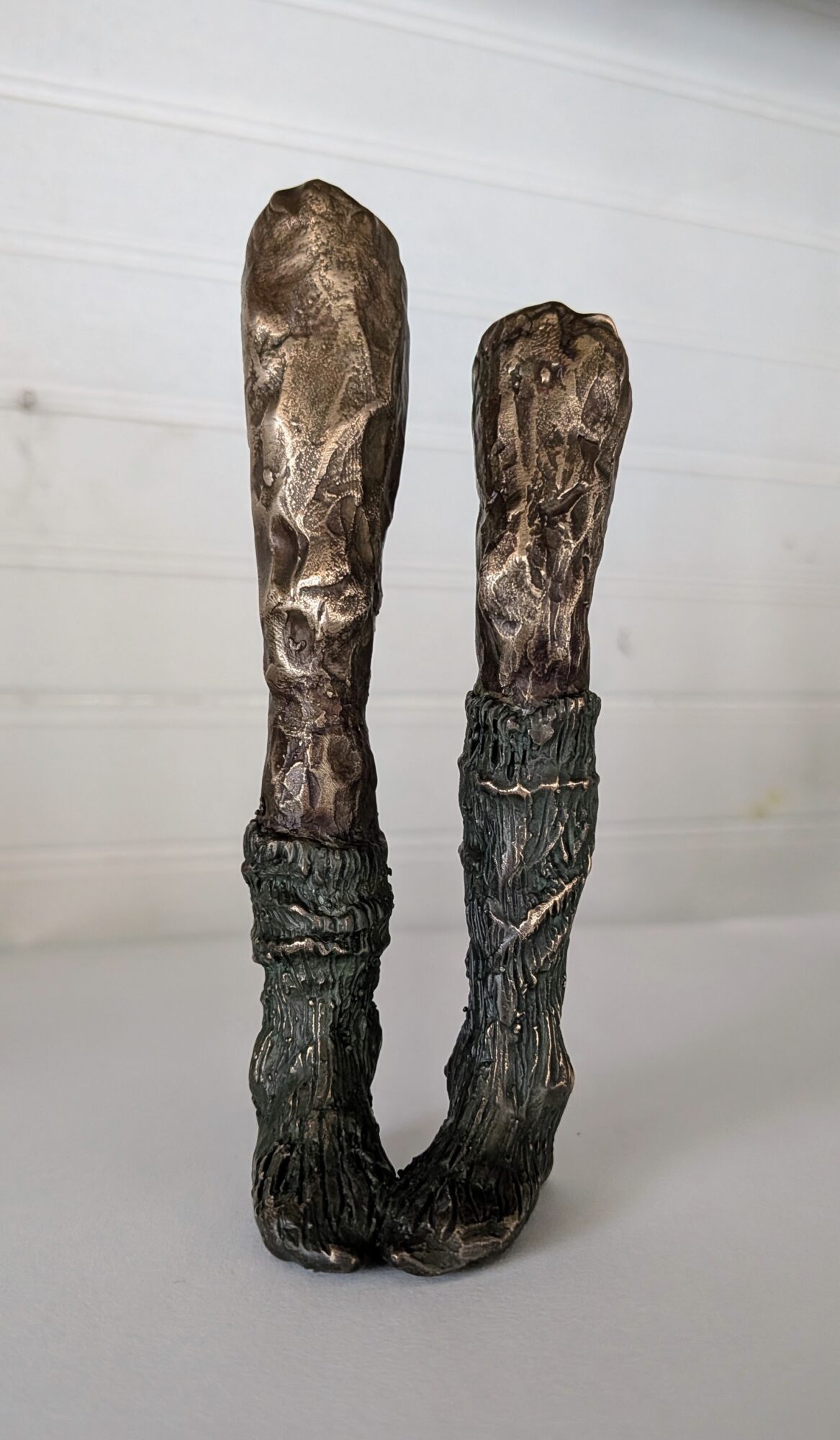 Two tall, textured wooden sculptures resembling legs or boots, standing on a reflective surface against a plain background.