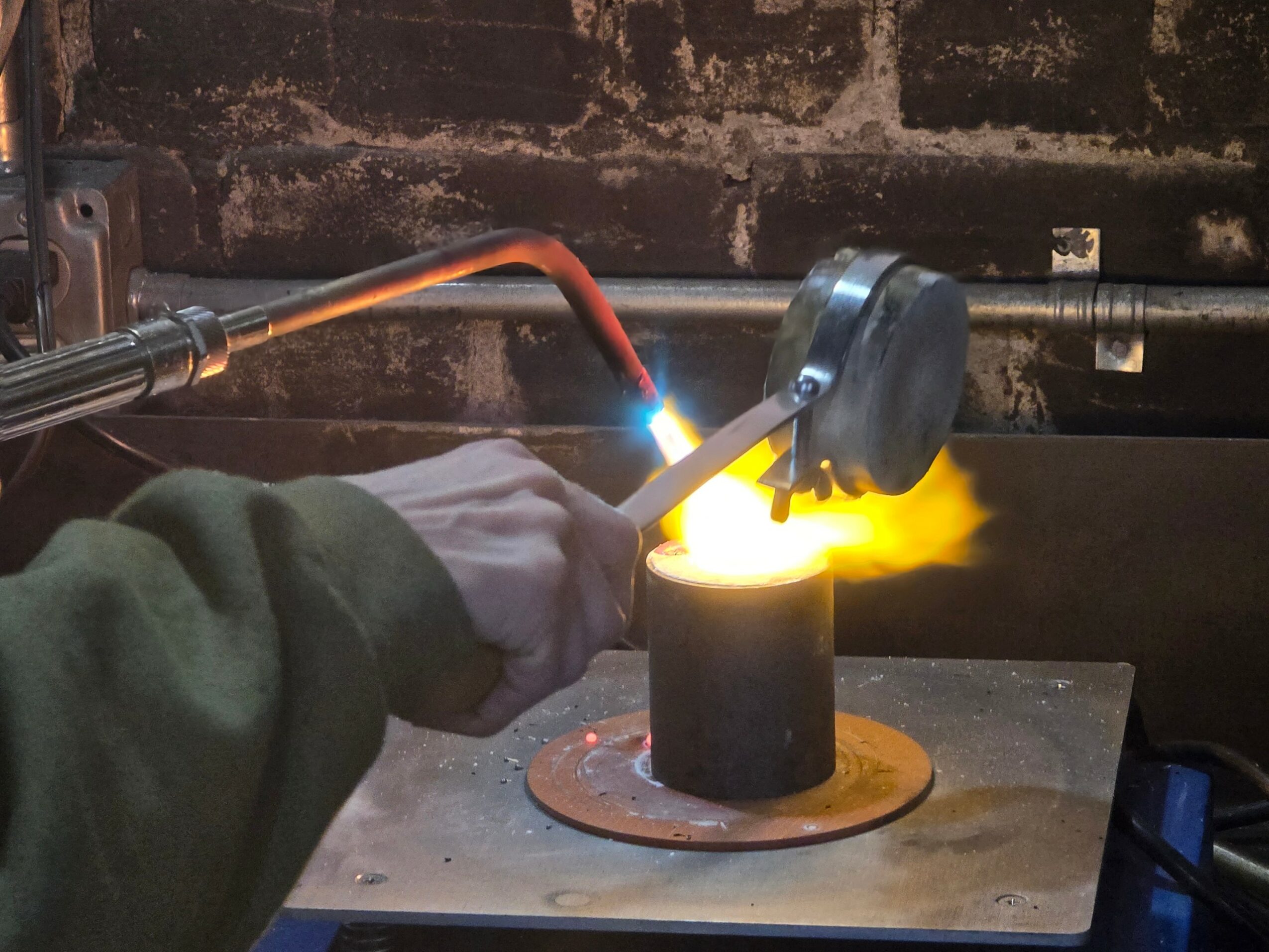 Person welding metal cylinder with bright sparks and flame, wearing a green sleeve, in a workshop setting.