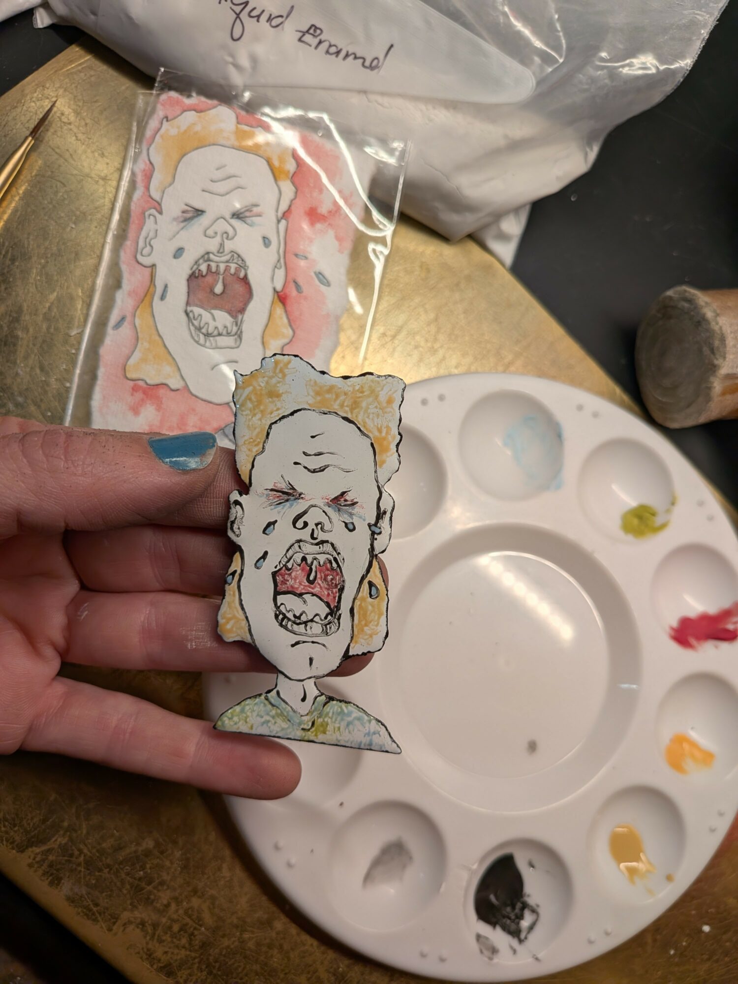 Two hand-painted zombie face pins with open mouths and rotting features, held over a paint palette and workspace.