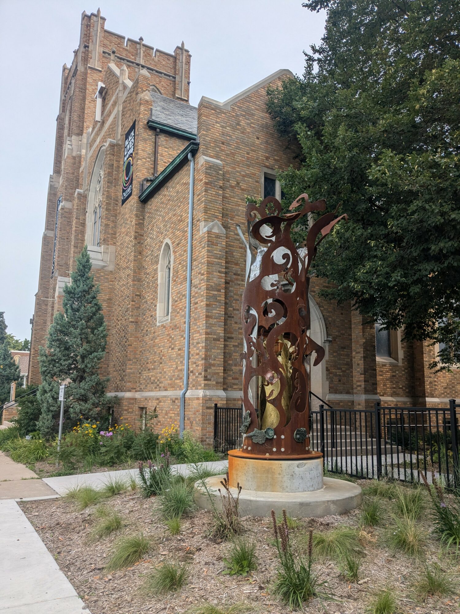 Brick church with tall tower, trees, and a metal sculpture in front, on a landscaped area.