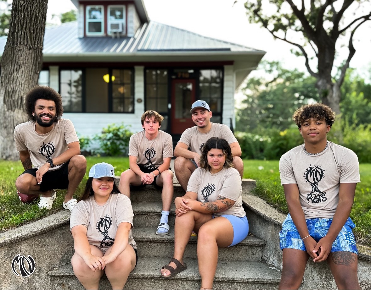 Group of six people sitting on steps outside a house, smiling, with trees and a house in background.