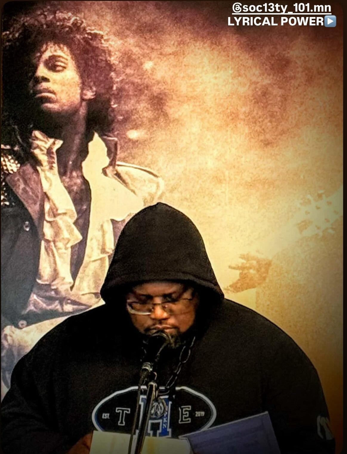 A person wearing a black hoodie and glasses, looking down, with a large poster of a woman with curly hair in the background.