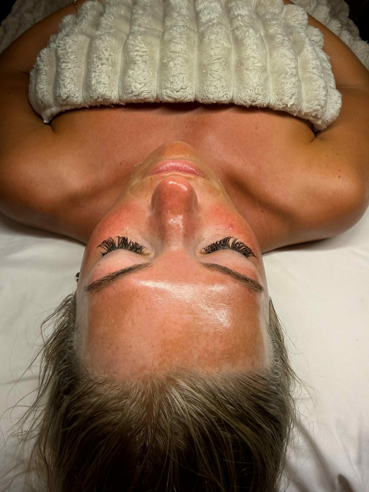 Woman lying down with eyes closed, hair spread out, wearing a white textured top, skin glistening.
