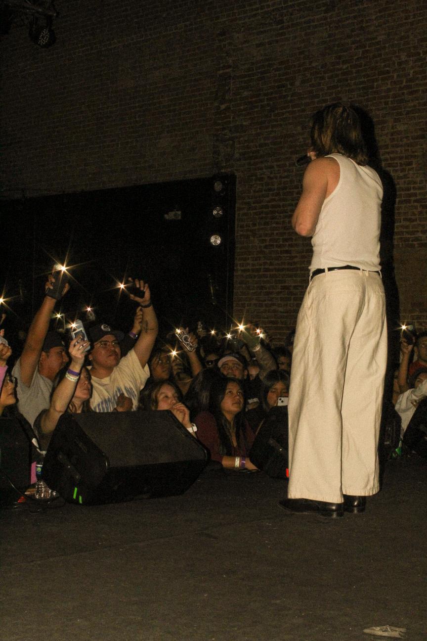 Person on stage facing audience, audience members holding up lights, dark background, brick wall, night setting