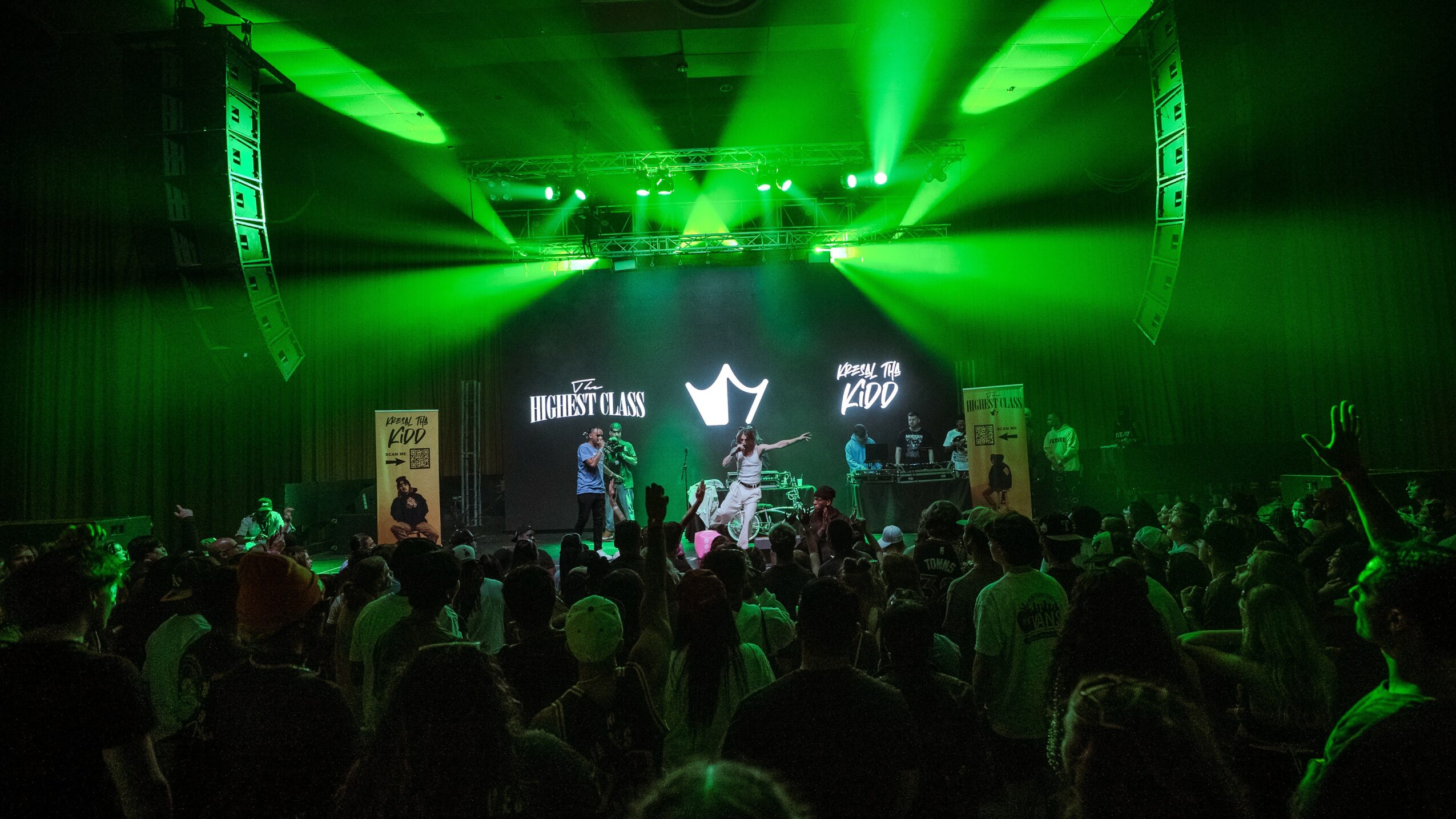 Concert scene with green stage lights, audience, and performers on stage, with a large screen displaying graphics.
