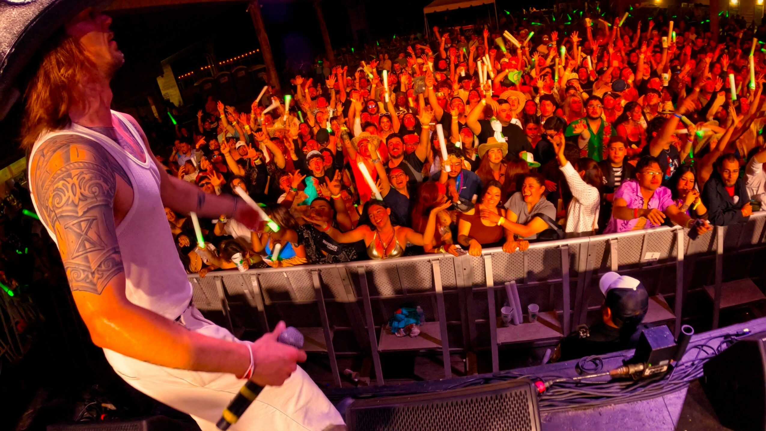 Performer on stage with a large crowd raising hands at a concert, colorful lighting, and stage equipment visible.